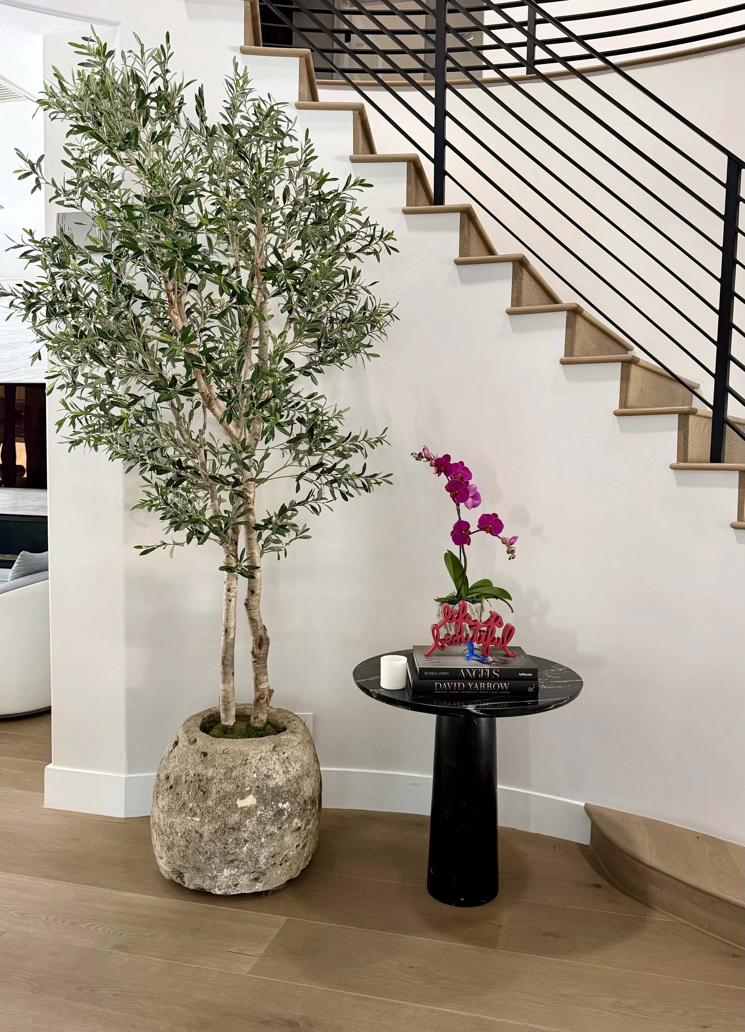 Interior view of a modern living space featuring a large potted olive tree, a small black side table with books and decorative items, and a staircase with wooden steps and black metal railing.