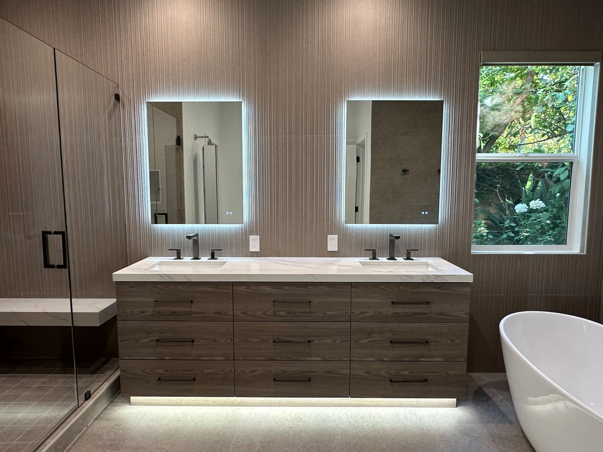Modern bathroom with two mirrors backlit with LED lighting, a double sink vanity with wood cabinetry, a window with greenery outside, and a bathtub with a curved shape.
