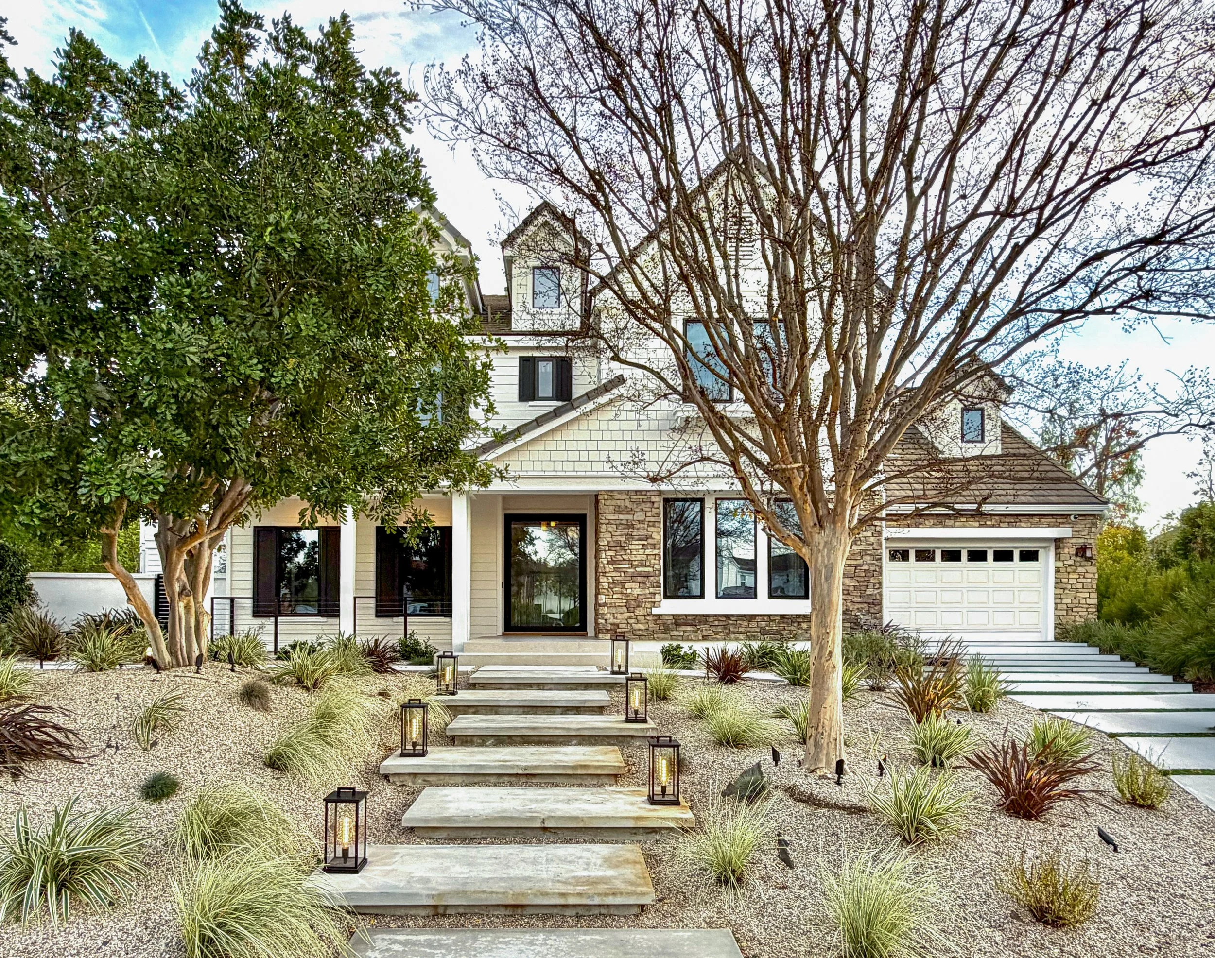 Front yard view of a modern house with a stone and siding exterior, a pathway with lantern lights, leafless and green trees, and a clear sky.
