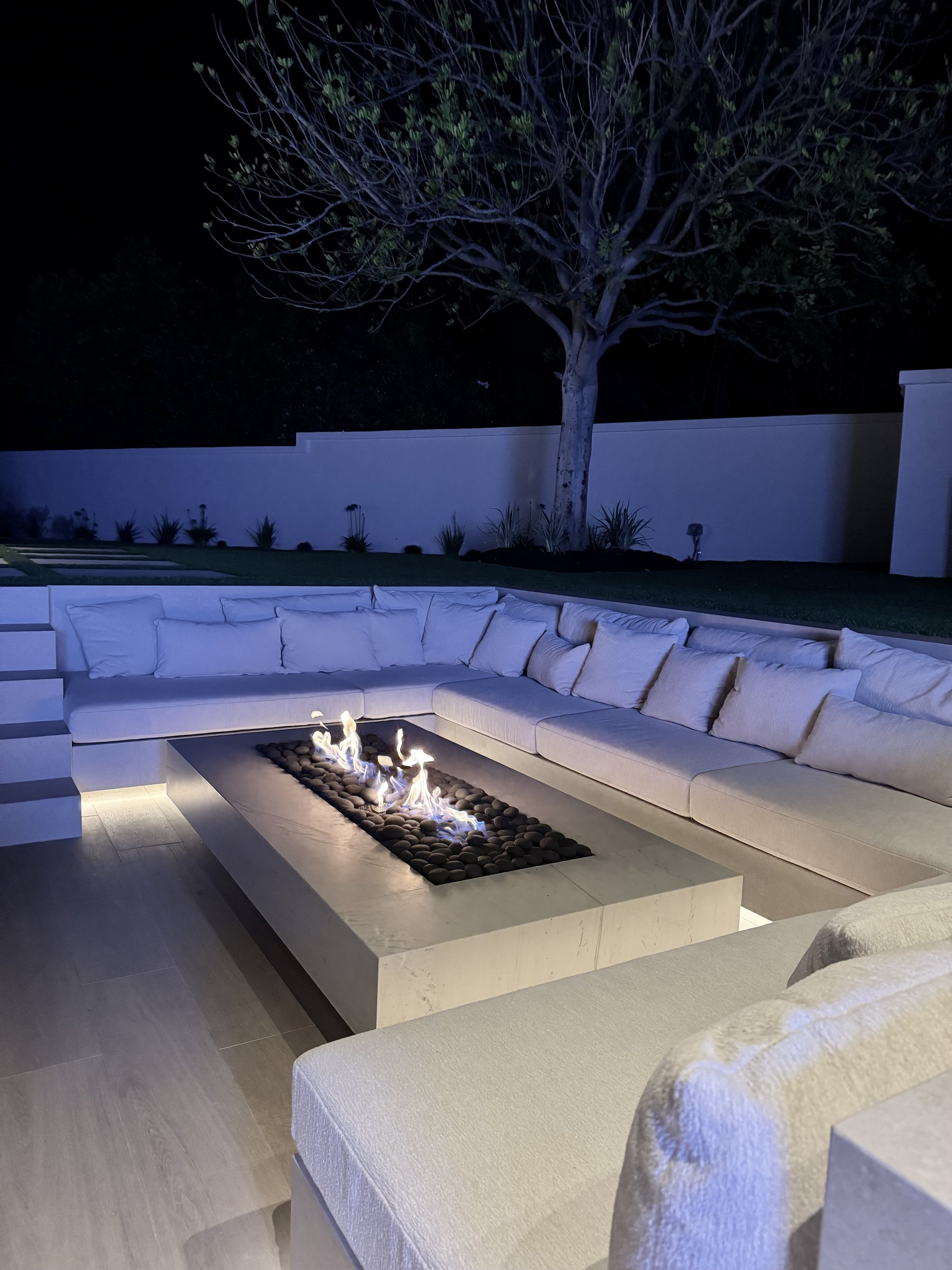 Nighttime outdoor patio with a modern rectangular fire pit surrounded by cushioned seating, a large tree, and a white privacy fence in the background.