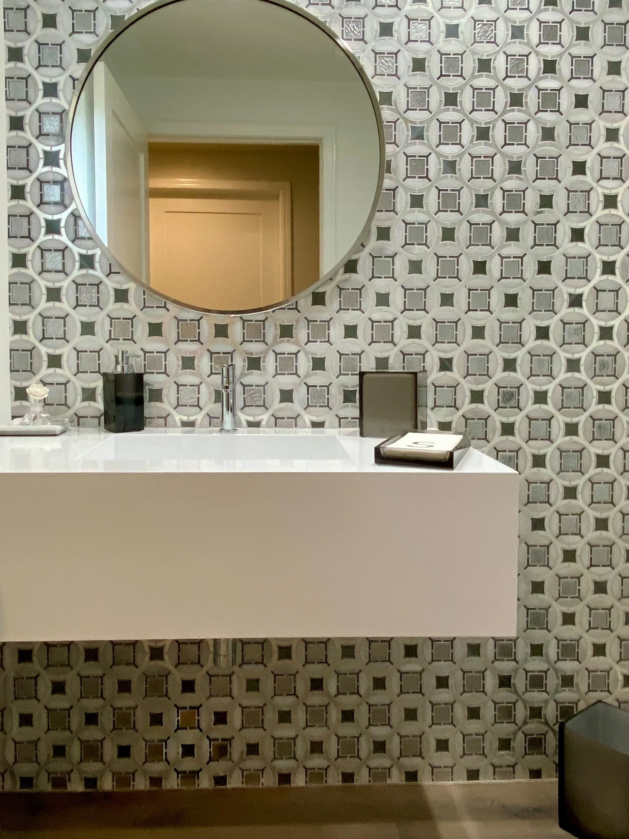 A bathroom sink area with patterned wall tiles, a round mirror, a soap dispenser, a tissue box, and a small vase with white flowers on the countertop.