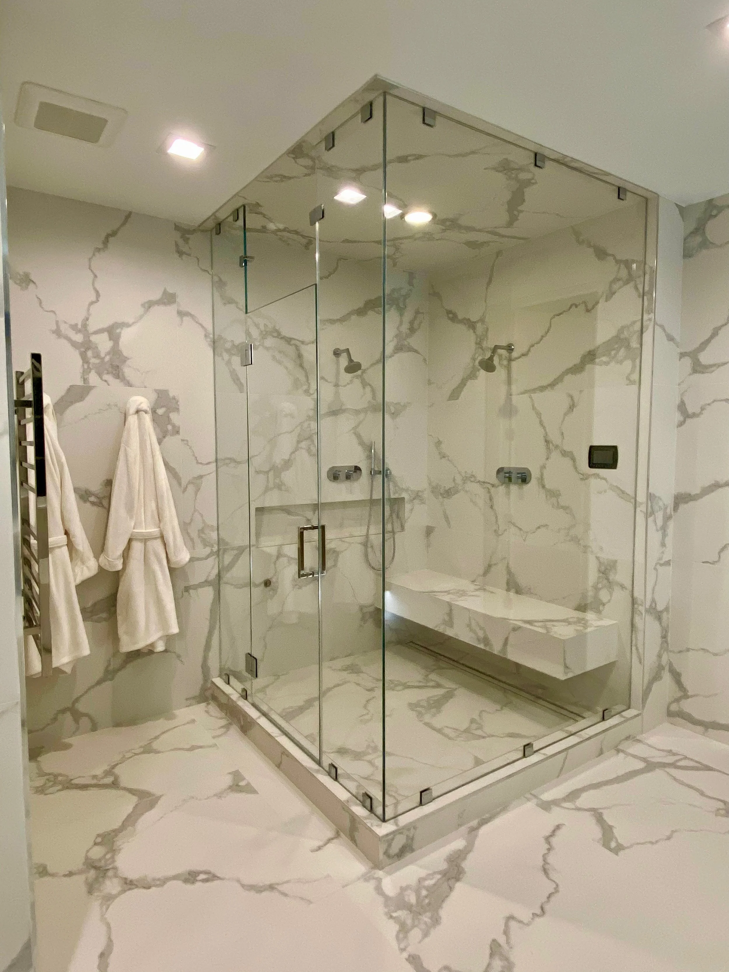A bathroom with marble walls and floors, and a glass shower enclosure with built-in seating and dual shower heads.