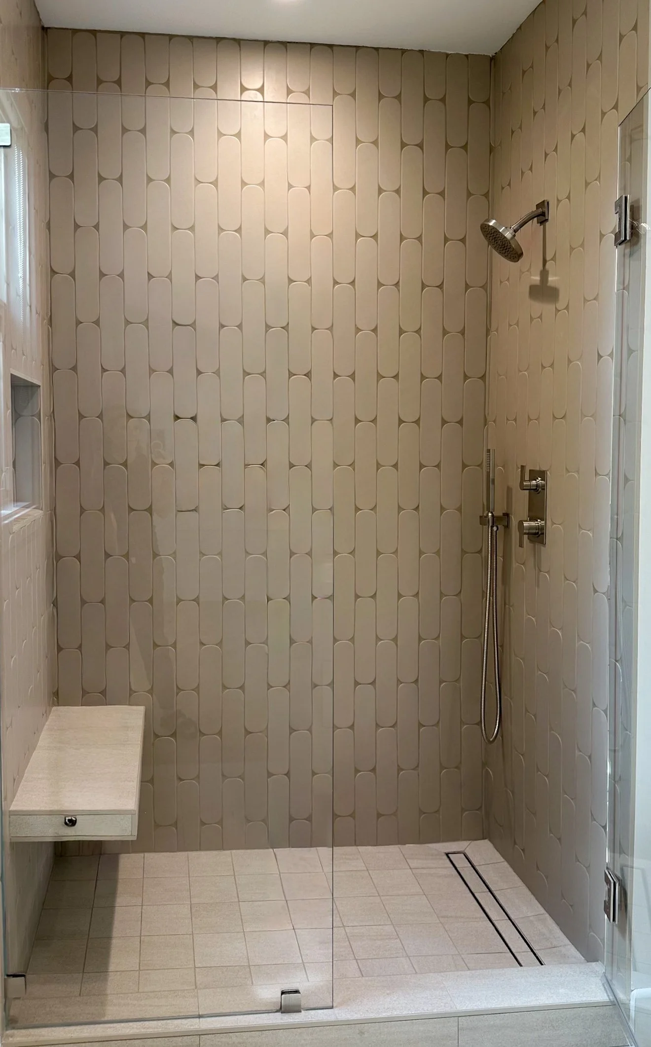 A modern shower with a beige and white patterned tile wall, a rain showerhead, a handheld shower, and a small bench on the left side.