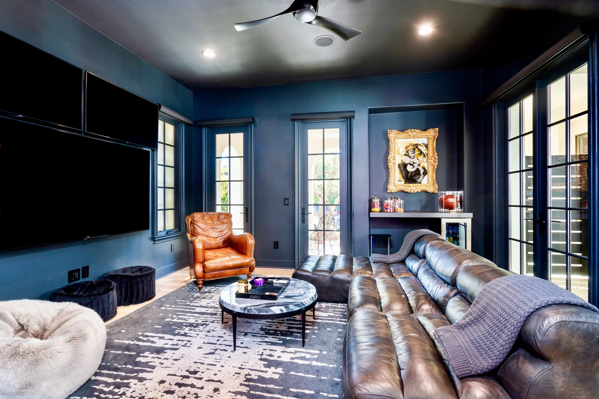 Living room with blue walls, large windows, two black flat-screen TVs, a leather sectional sofa, a round marble coffee table, a leather armchair, and various decorative items including framed artwork.