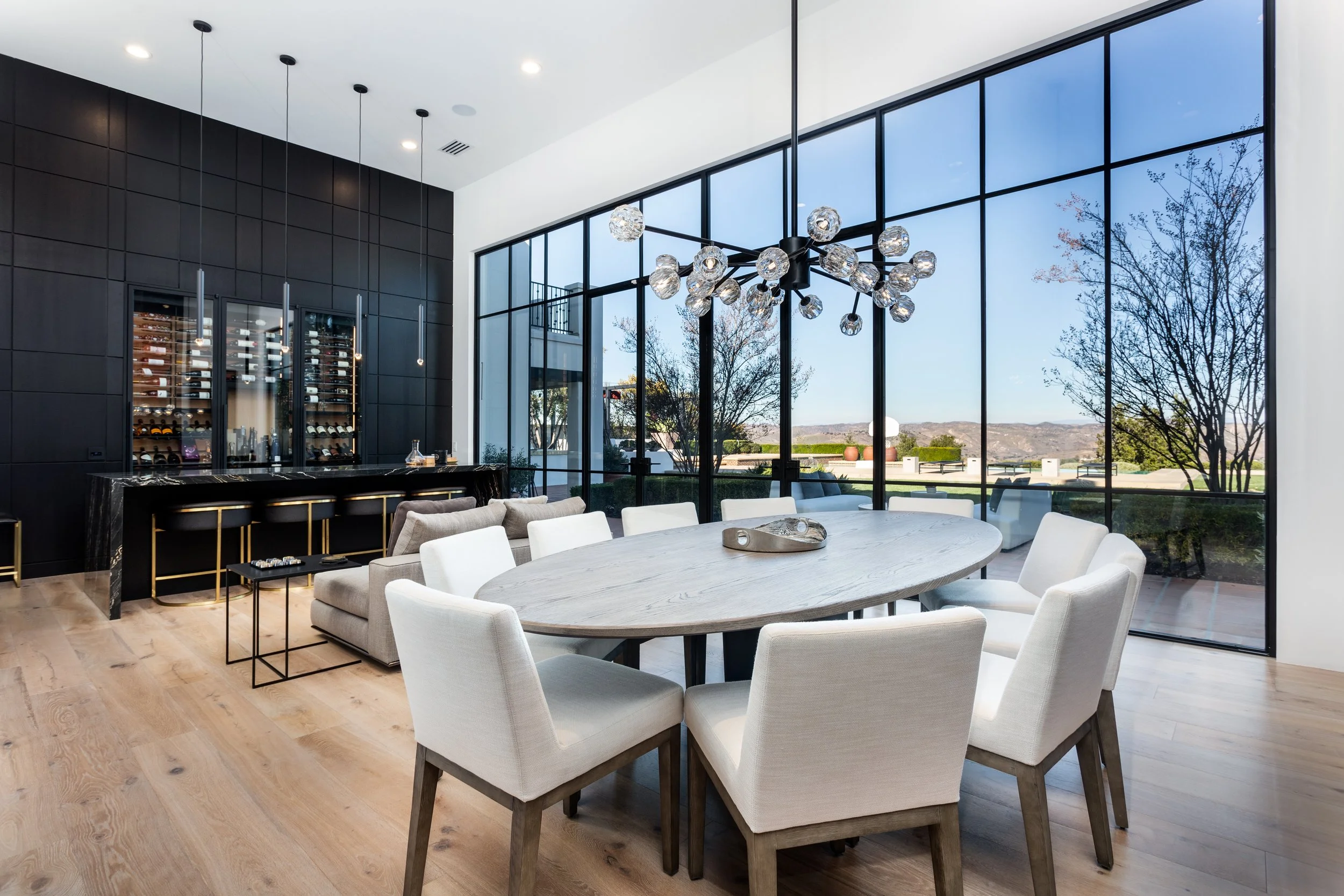 Modern dining room with large glass windows, white upholstered chairs around a round table, black pendant lights, and a black wine fridge built into a dark wood wall.