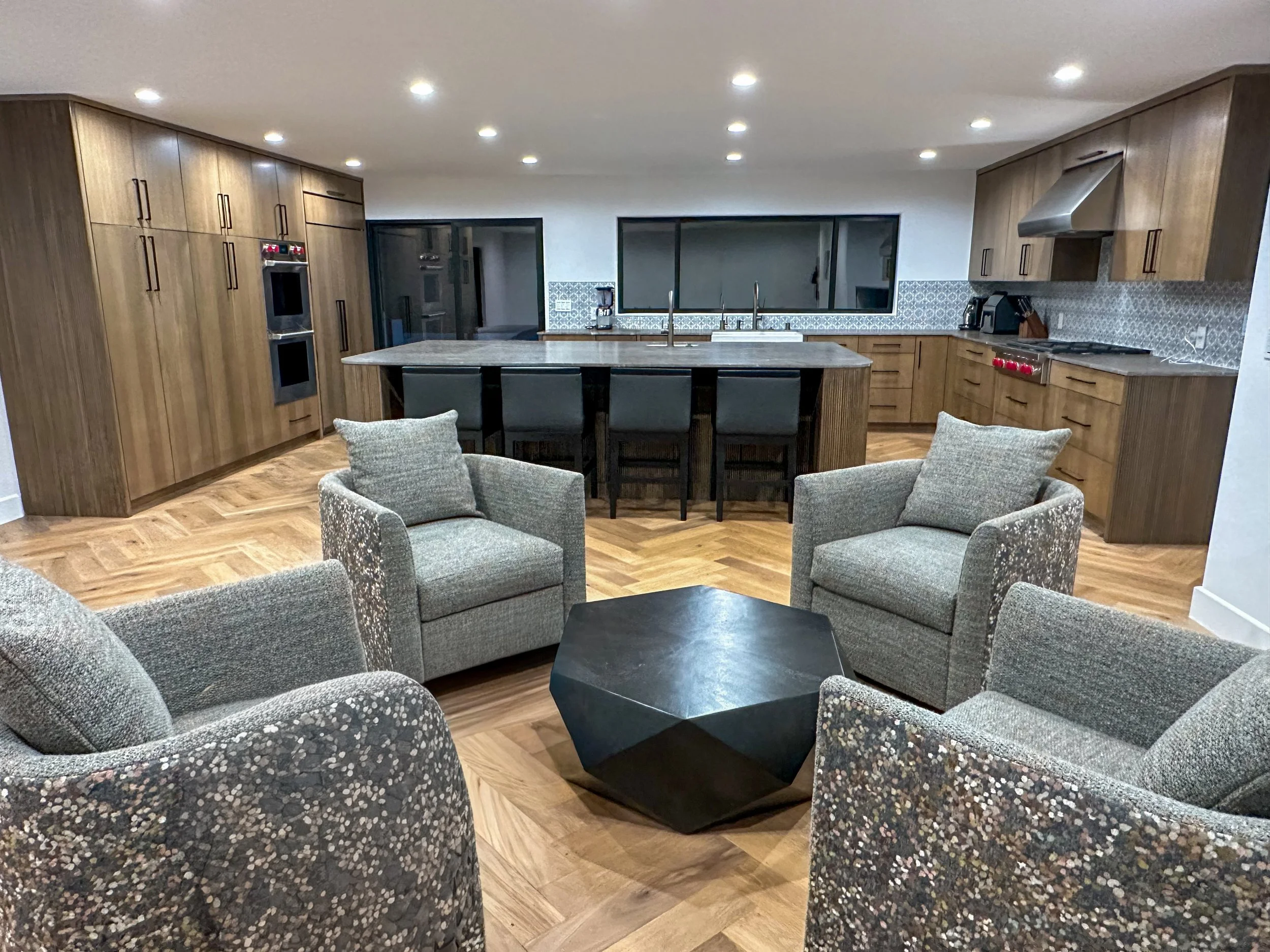 Living room with modern furniture, including a sectional sofa with textured fabric, black coffee table, and wood flooring, leading into a kitchen with wooden cabinets, granite countertop, and stainless steel appliances, illuminated by ceiling lights.