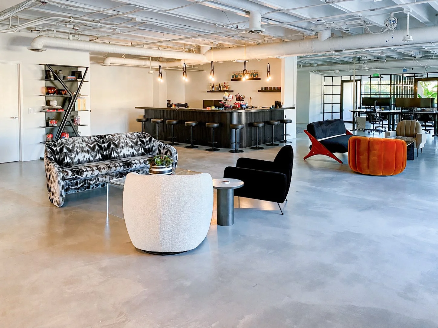 Modern open-concept office lounge with assorted seating, a bar area with stools, and workstations in the background. Decor includes a black-and-white patterned sofa, colorful lounge chairs, and shelving with books and decor items.