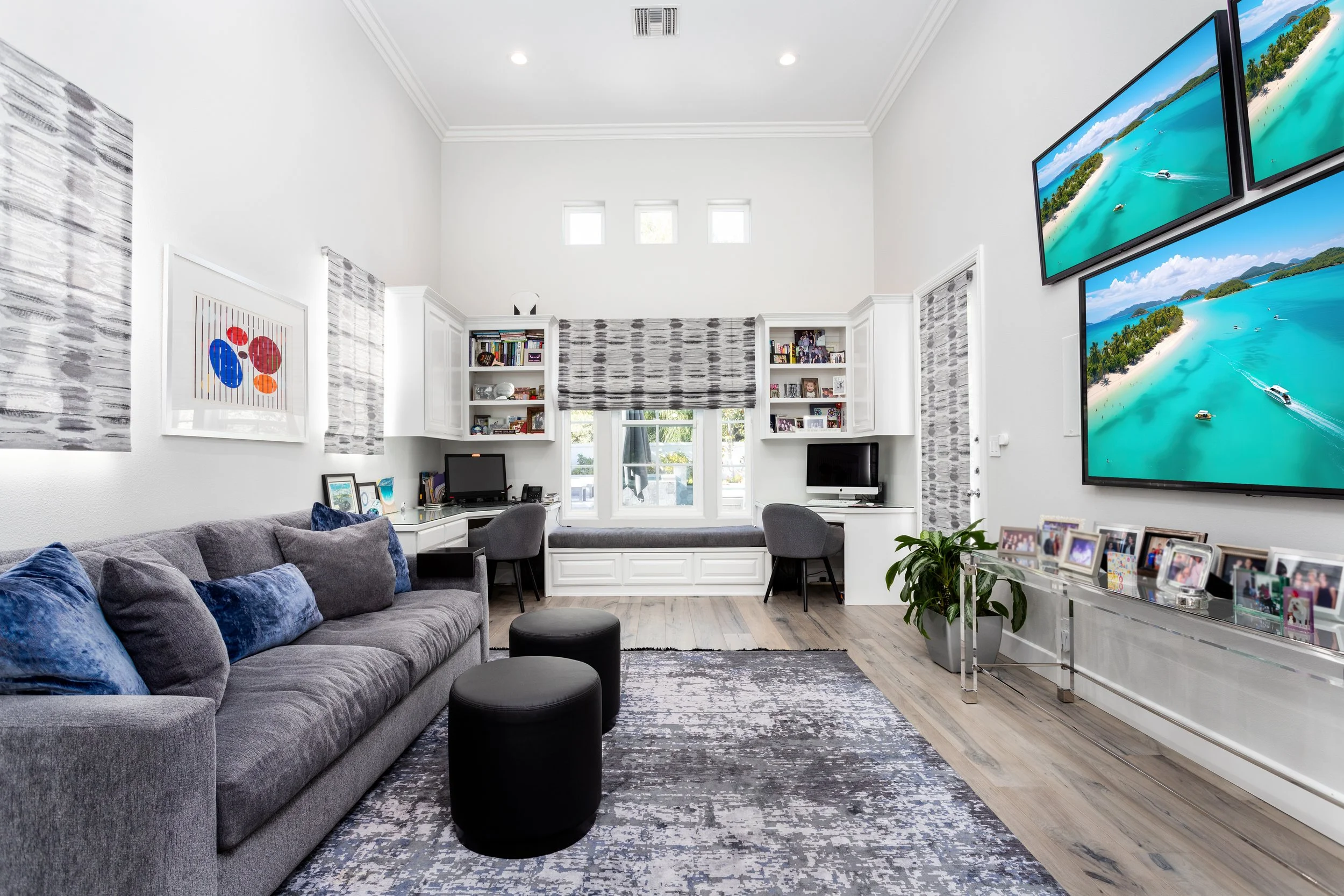 A living room with a large gray sofa with blue pillows, a black ottoman, a patterned rug, white built-in shelves with books and photos, a window seat with cushions, three small square windows high on the wall, and three large screens displaying beach