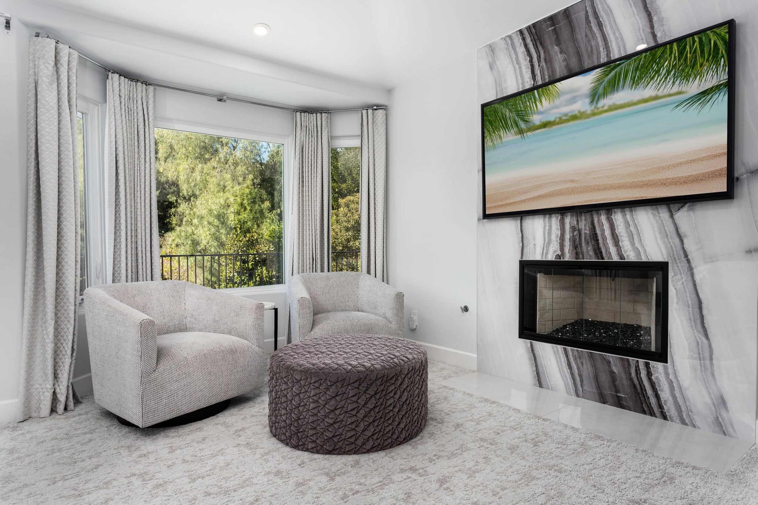 Modern living room with white walls, a large window with patterned curtains, a wall-mounted flat-screen TV displaying a beach scene, a fireplace, beige upholstered chairs, and a textured ottoman.