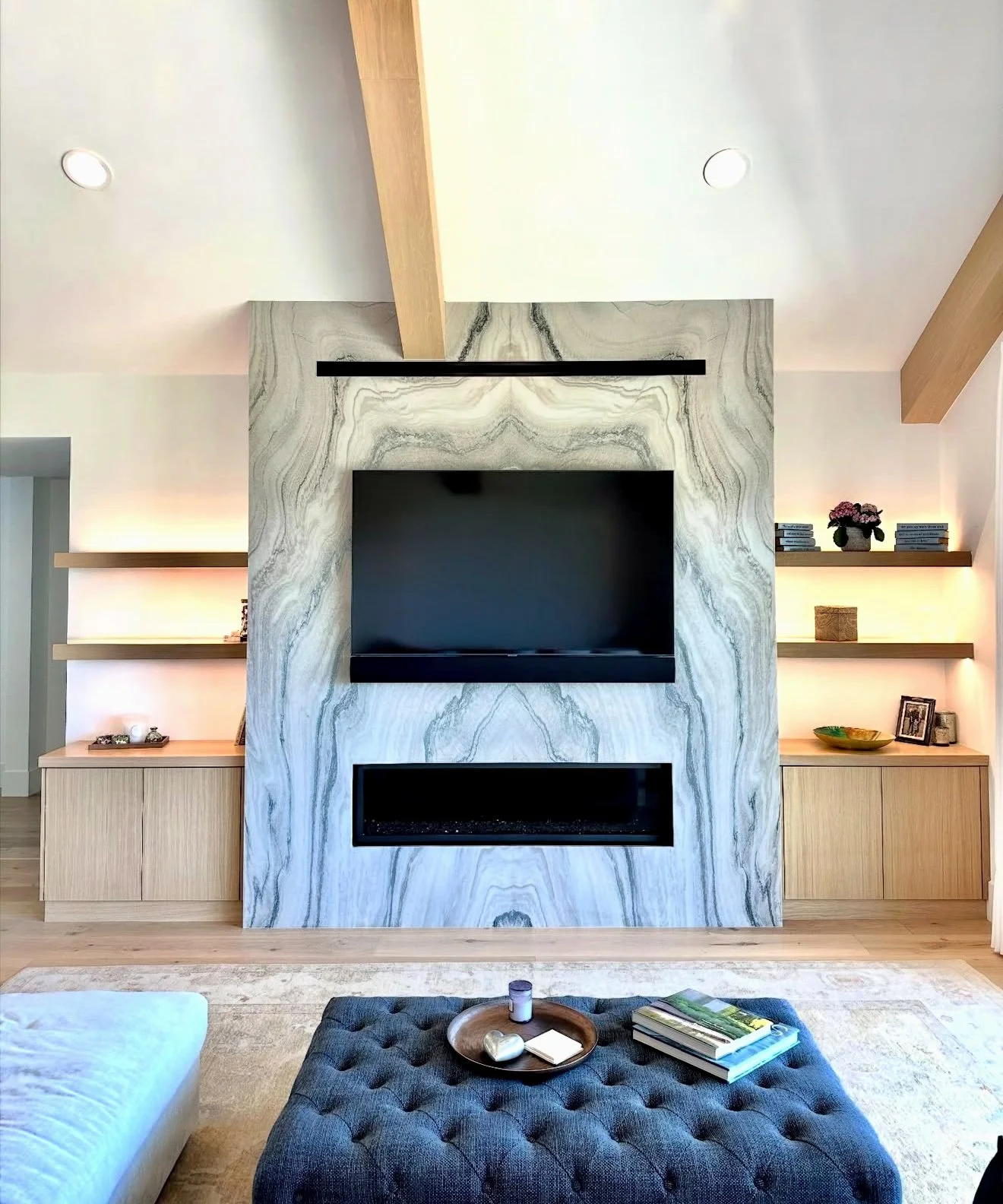 Living room with a central feature wall made of marble with gray and white veining, a wall-mounted flat-screen TV, and a modern fireplace below it. On either side, there are built-in wooden shelves with decorative items, plants, and books. In the for