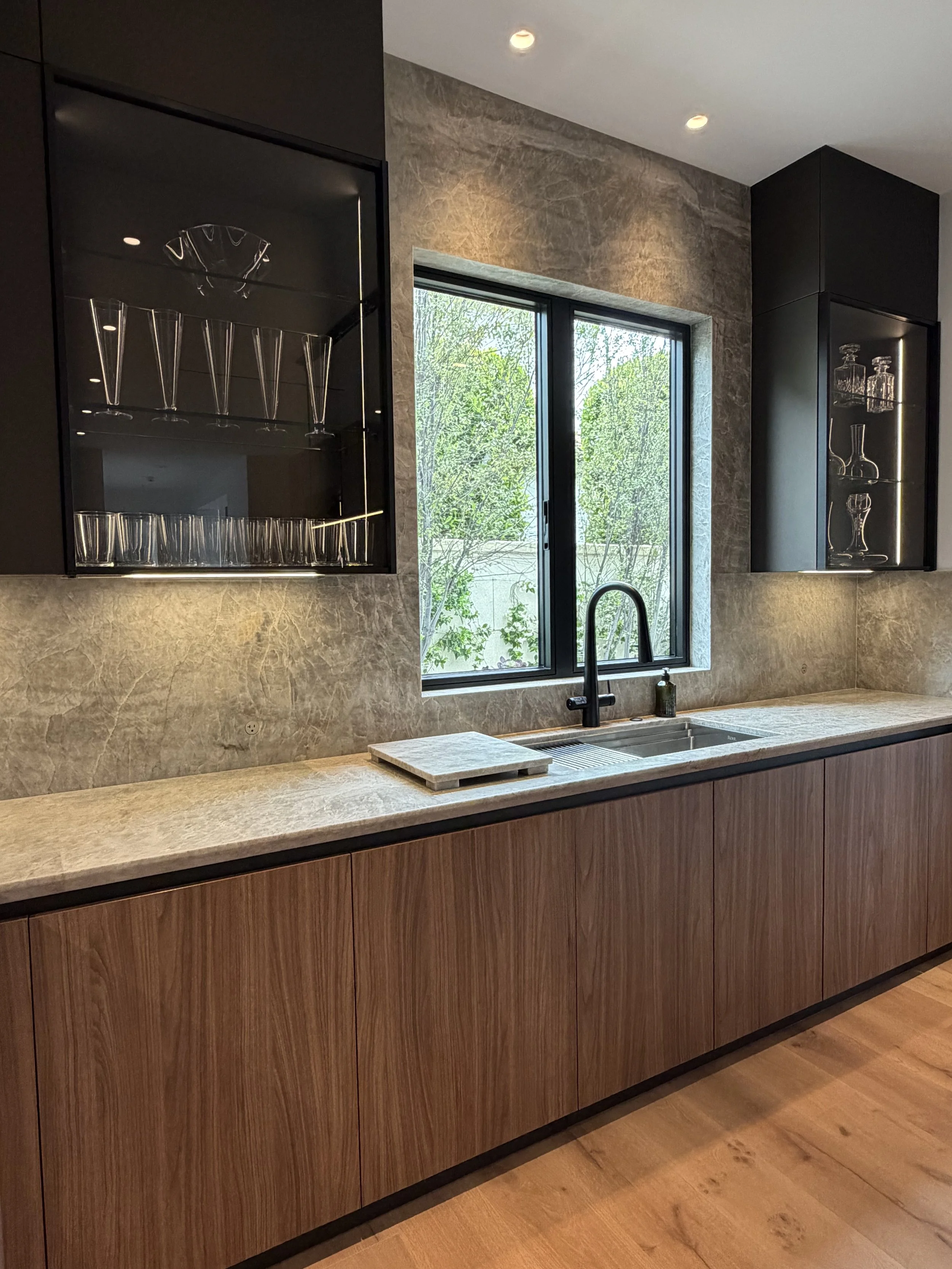 Modern kitchen with wooden cabinets, beige marble countertop, black sink faucet, window with a view of green trees, glass shelf with glasses and decanter, and warm ambient lighting.