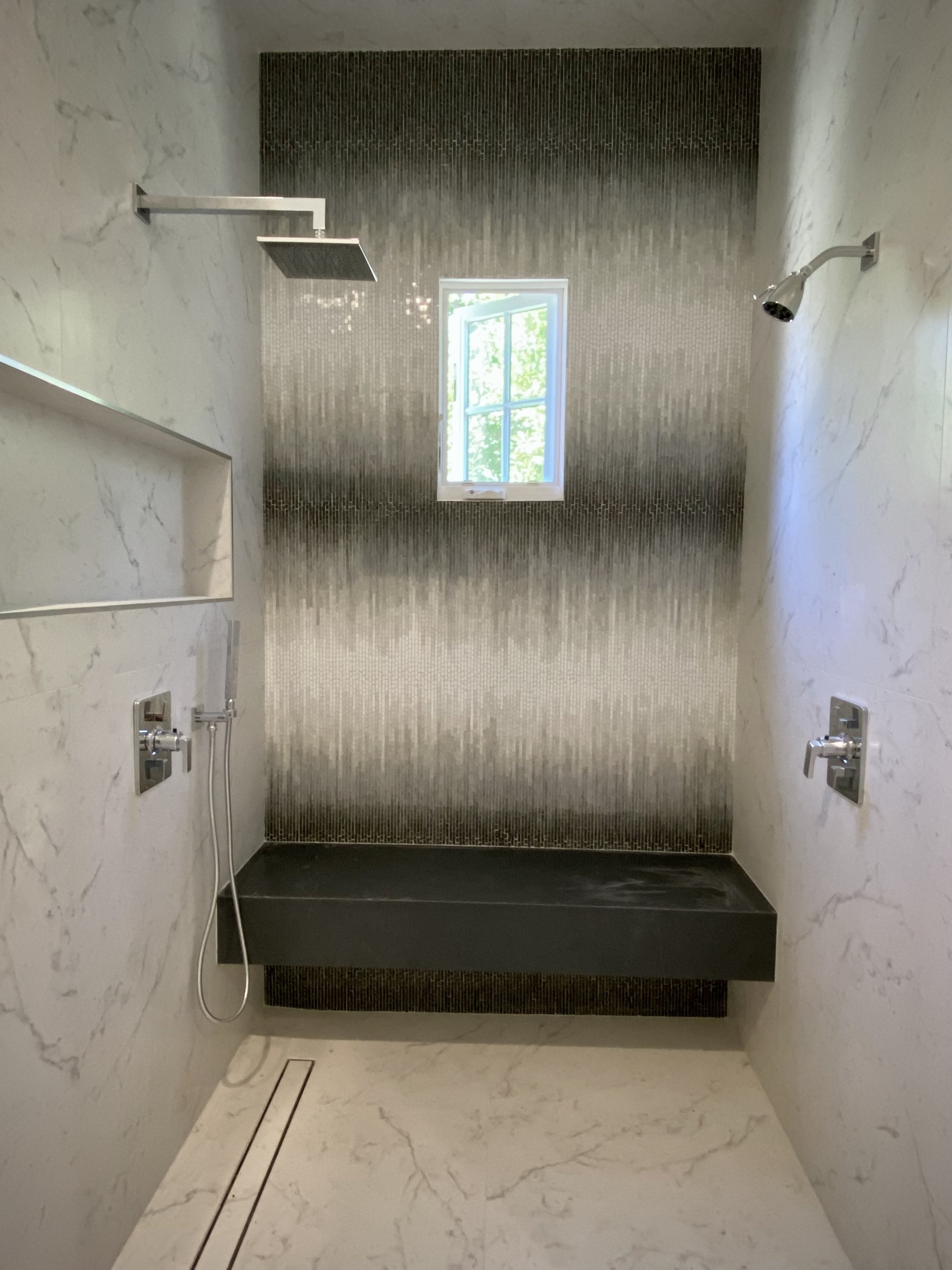 Modern shower area with marble walls, a small window, two shower heads, and a built-in seating area.