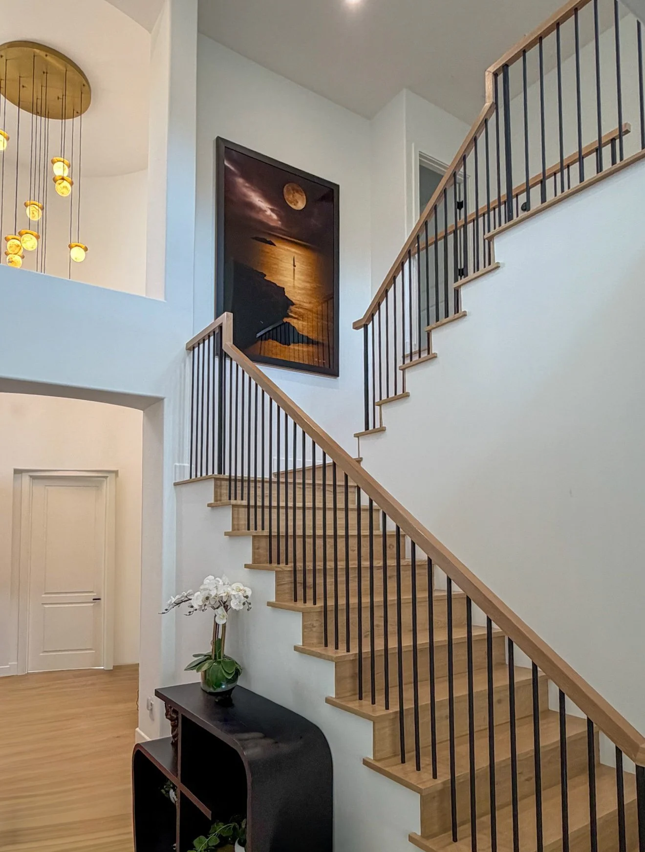 Interior view of a staircase with wooden steps, black metal railing, white walls, a potted orchid plant on a black table, a large framed moonlit seascape painting, and a modern hanging light fixture with small yellow lights.