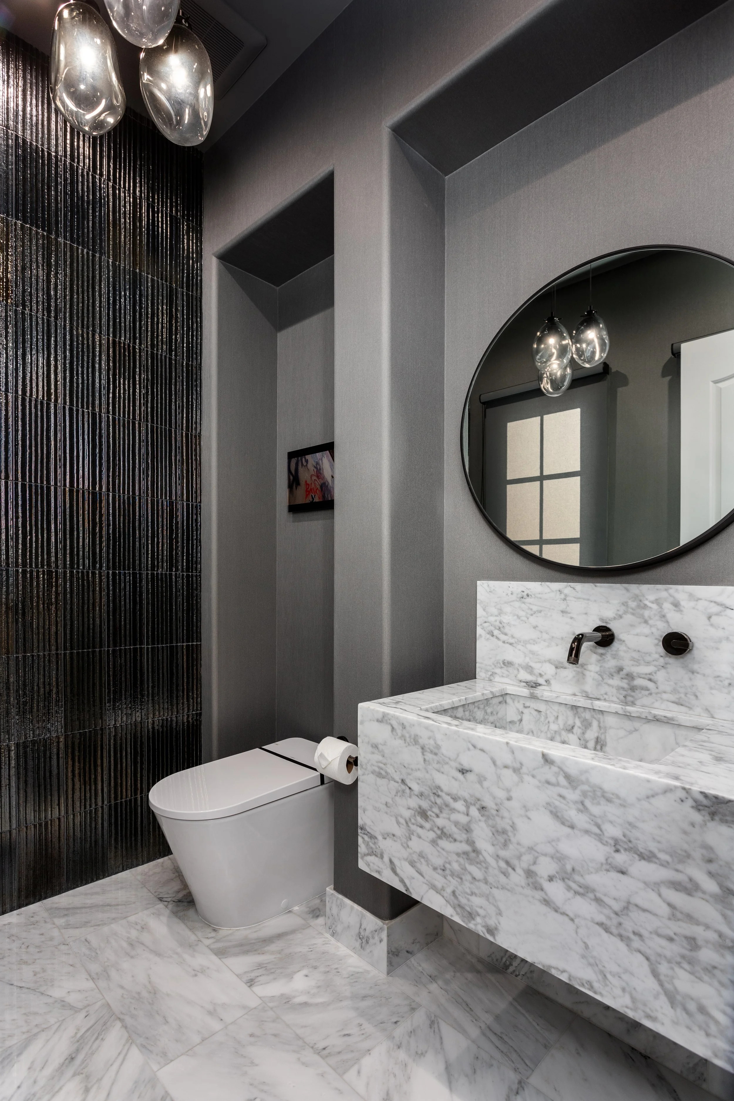 Modern bathroom with gray walls, black textured tile wall, oval mirror, pendant light fixtures, marble sink with a black faucet, and a wall-mounted toilet with a toilet paper roll