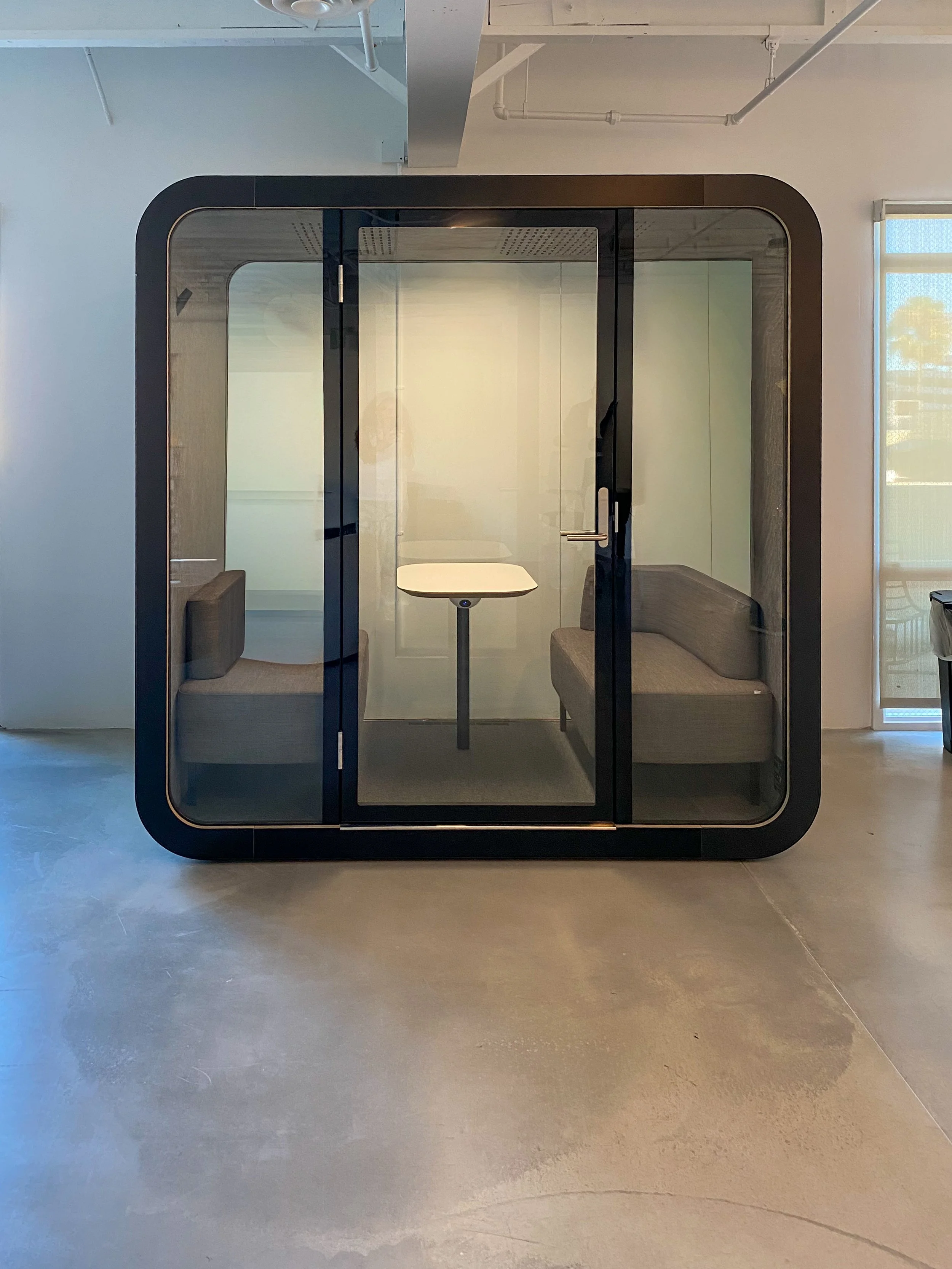A modern enclosed meeting pod with glass walls and rounded wooden edges, featuring built-in seating on either side and a small table in the center, situated in a bright, industrial-style room.