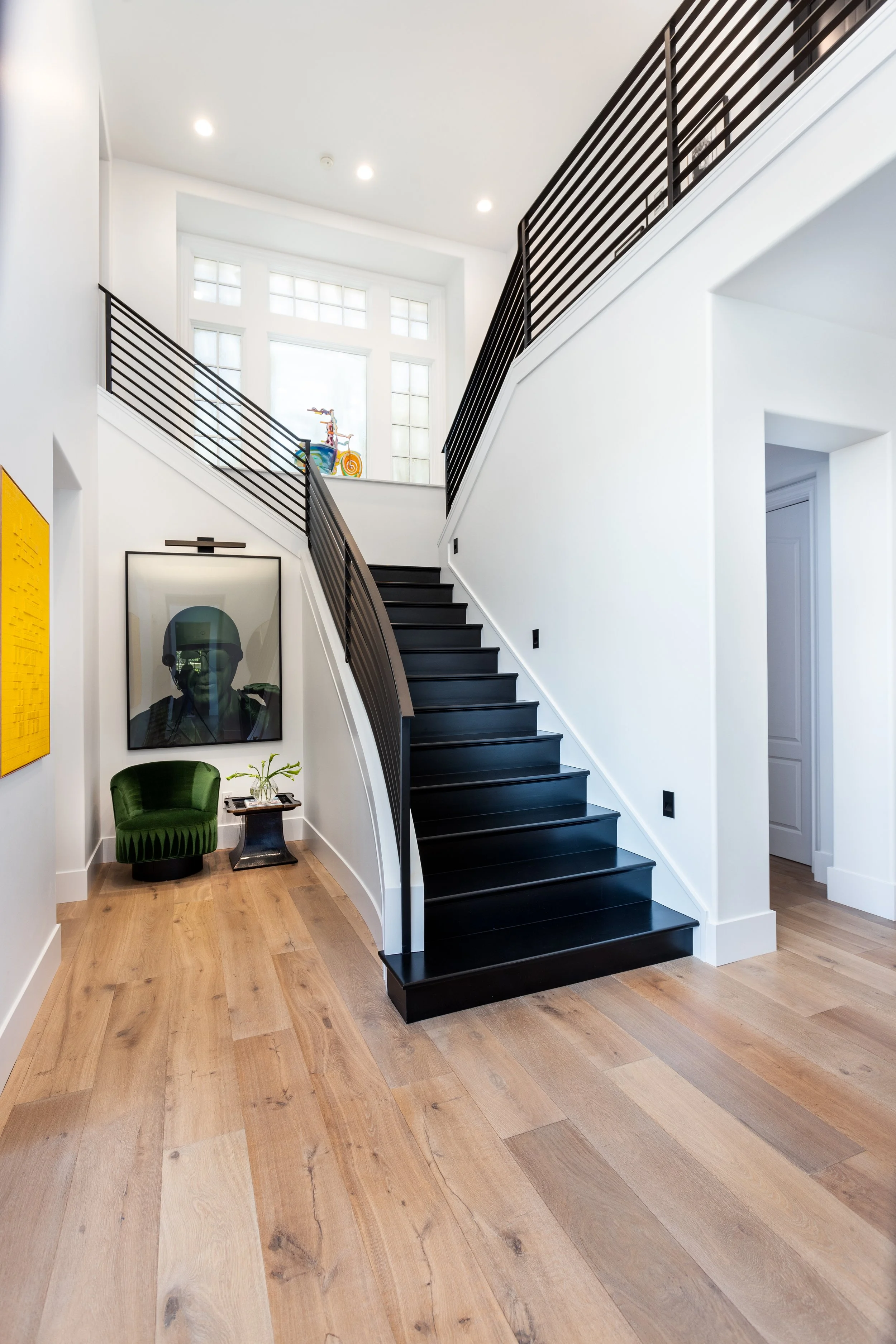 Interior of a modern house with a black staircase, light wooden flooring, and white walls. Decorative art pieces include a framed portrait and a yellow artwork on the wall, along with a green velvet chair and small side table.