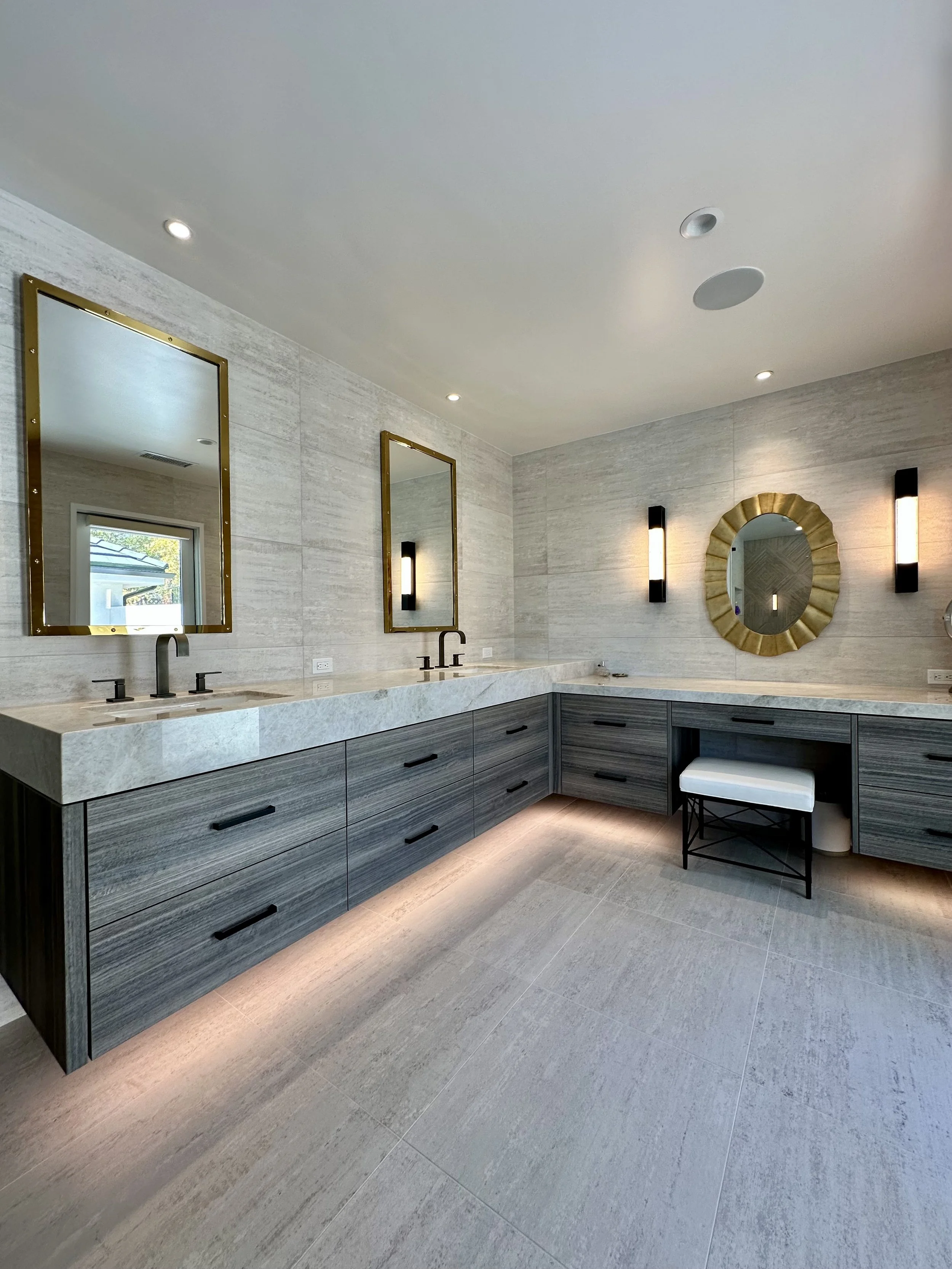 Modern bathroom with gray wooden cabinets, stone countertop, black fixtures, mirrors with gold frames, and decorative circular mirror on light gray tiled walls. Lighting includes wall sconces and ceiling spotlights.