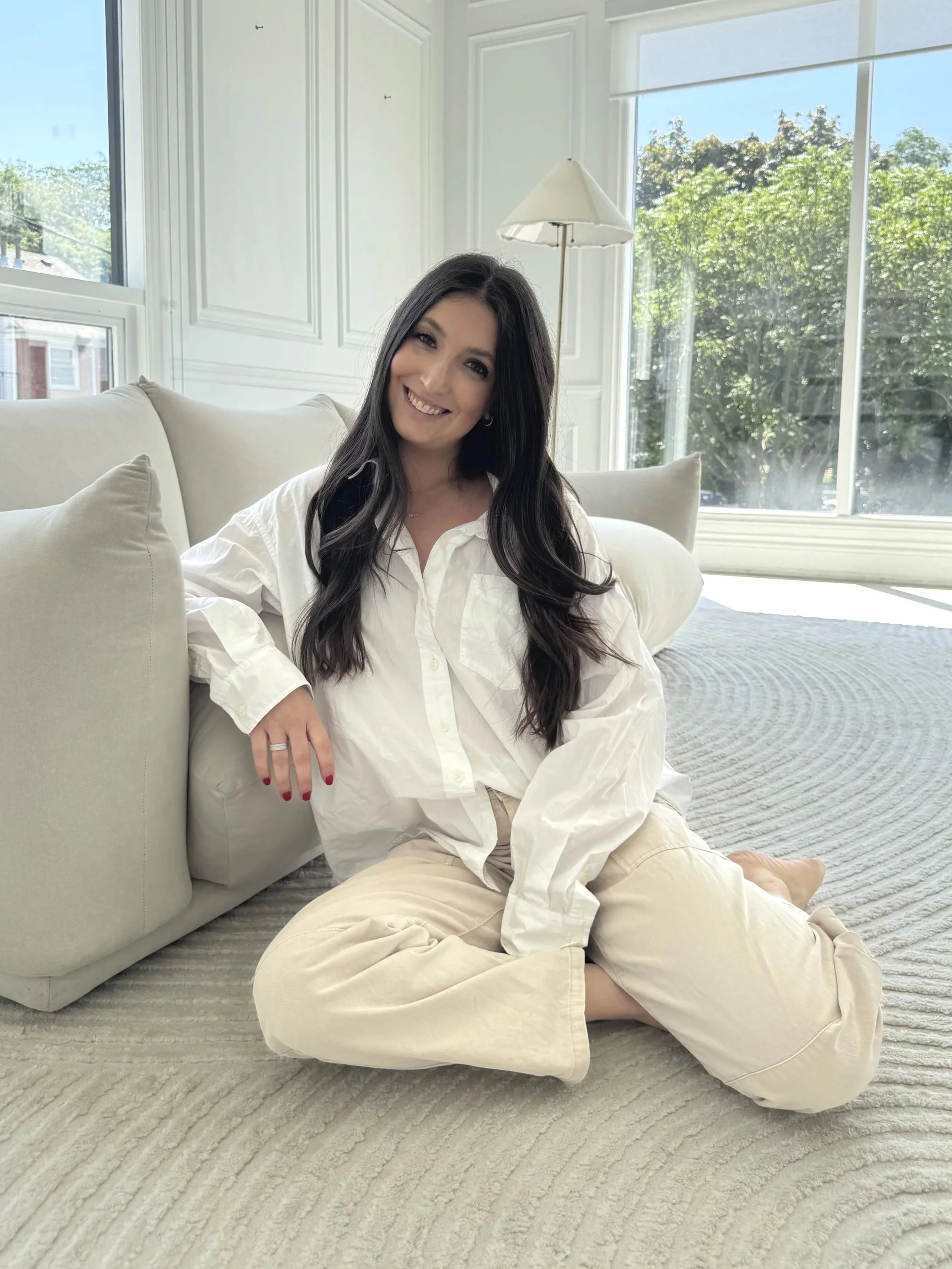 A woman with long dark hair smiling, sitting on a light-colored carpeted floor by a cream-colored sofa, in a bright room with large windows showing greenery outside.
