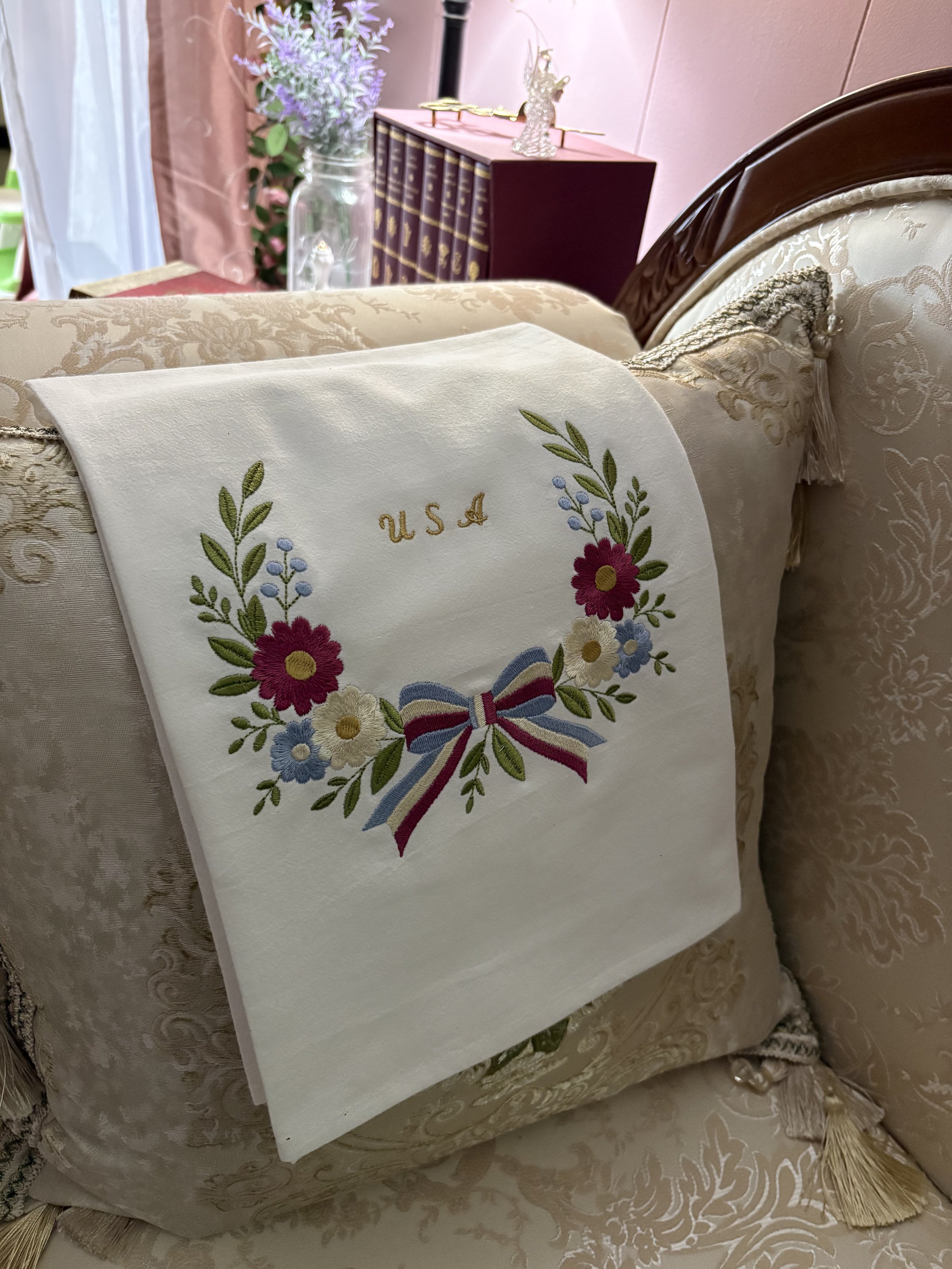 White flour sack tea towel featuring a patriotic wreath embroidered with red, white, and blue flowers, a ribbon, bow, and gold USA lettering in a vintage heirloom style.