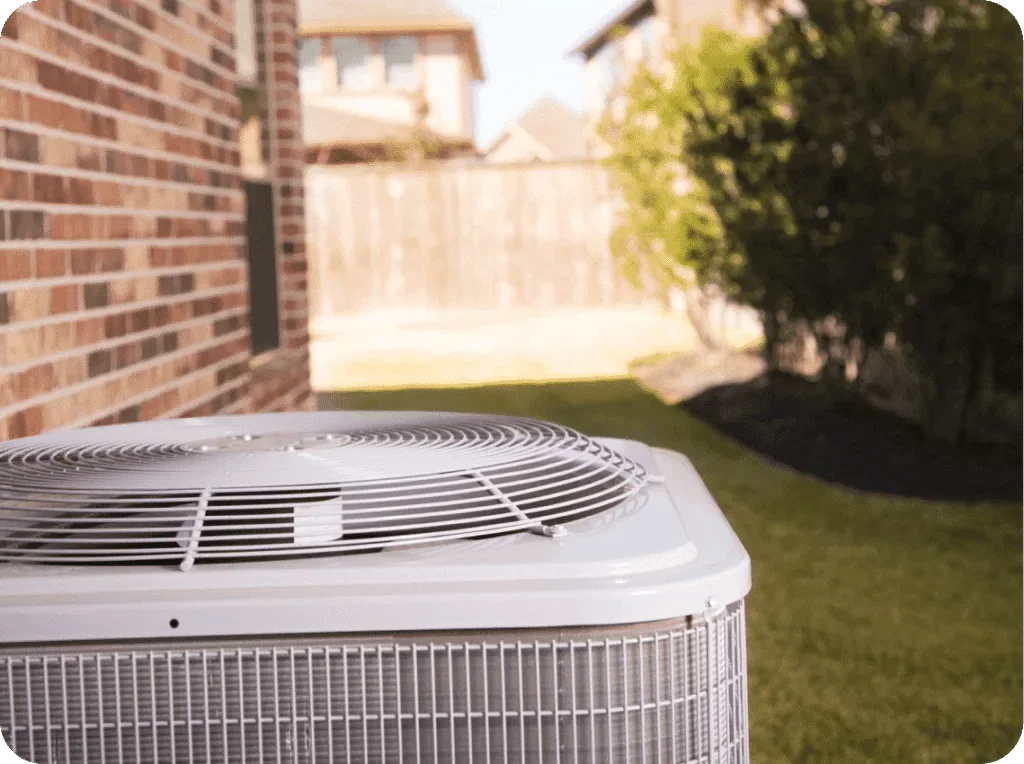 An air conditioning unit installed on a backyard lawn with a brick house on the left and bushes on the right, under a sunny sky.