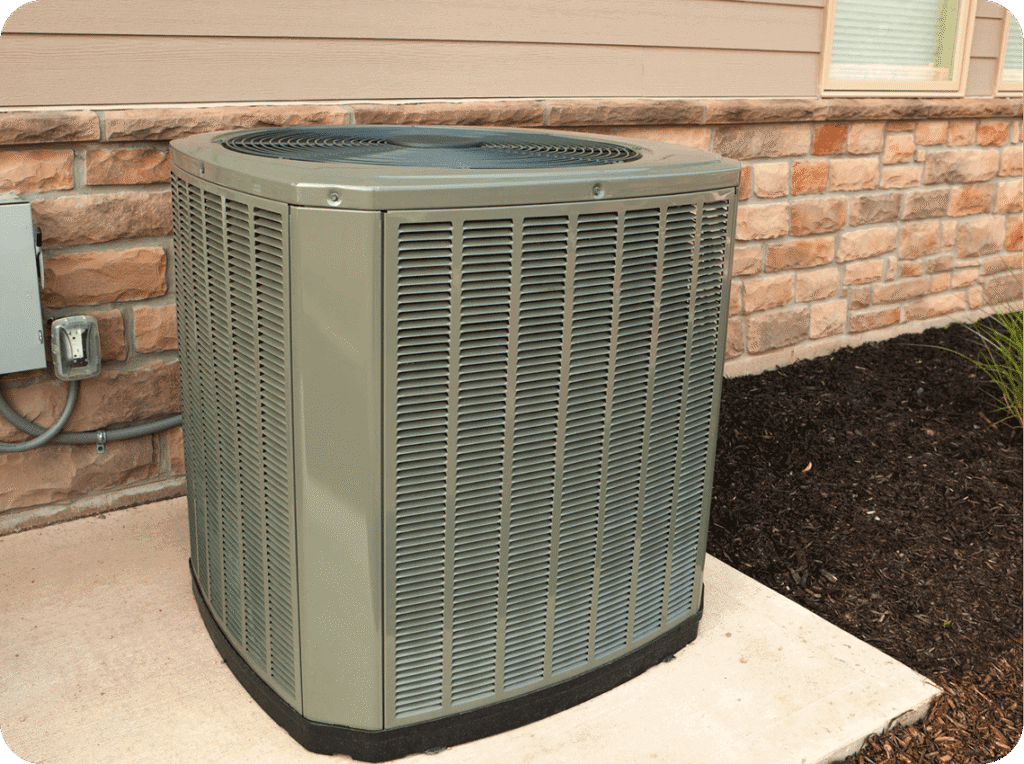 A green central air conditioning unit outside a house, situated on a concrete slab next to a brick wall and soil bed.