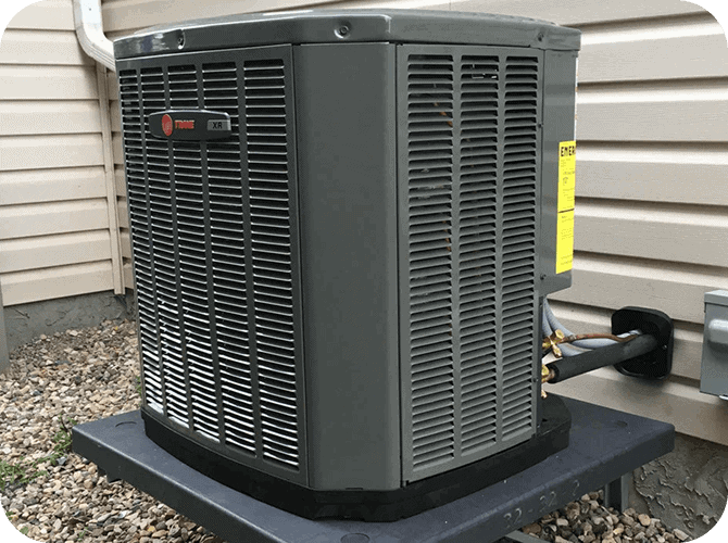 A large, gray central air conditioning unit installed outside on a black platform next to a house with beige siding.