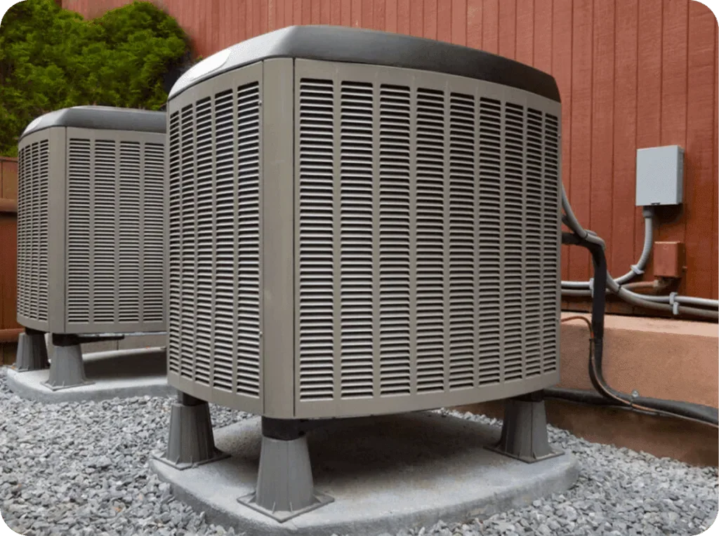 Two outdoor air conditioning units installed on a concrete pad, with a wooden fence and some greenery in the background.