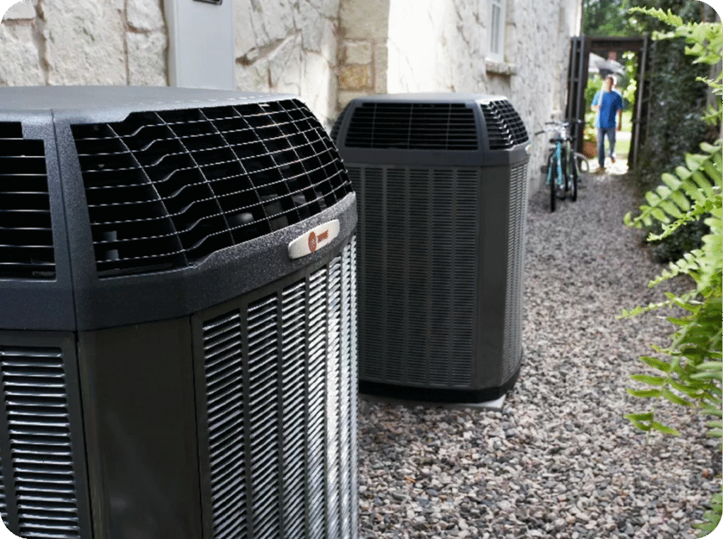 Outdoor scene featuring two large black air conditioning units on a gravel pathway next to a stone building with a garden gate and a person with a bicycle visible in the background.