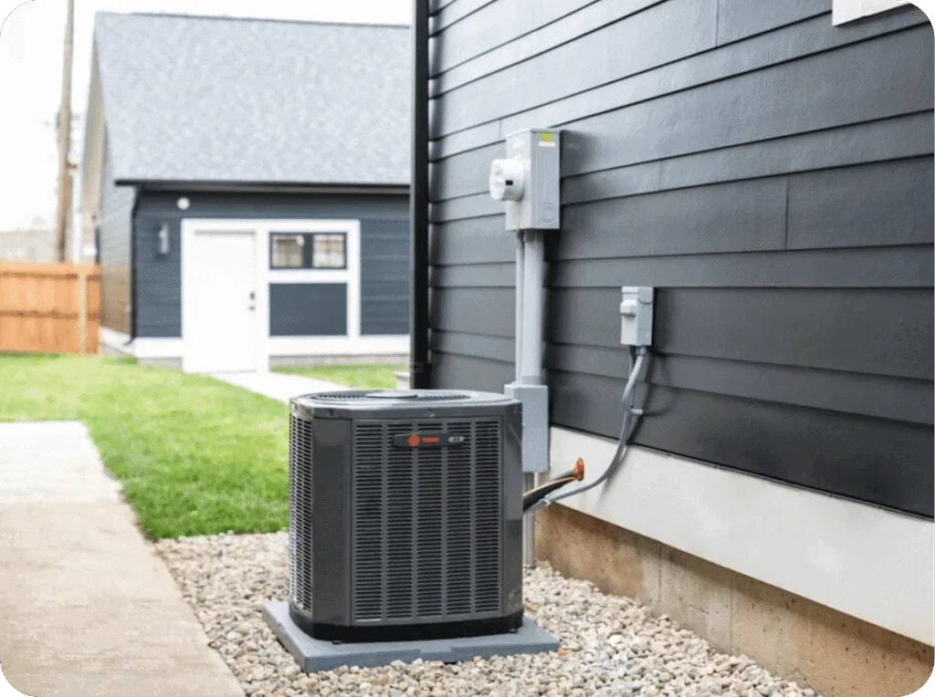 Outdoor air conditioning unit installed next to a black house siding, with electrical connections and a small shed in the background.
