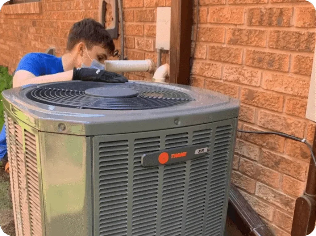 Person in blue shirt and face mask working on an outdoor HVAC unit against a brick wall.