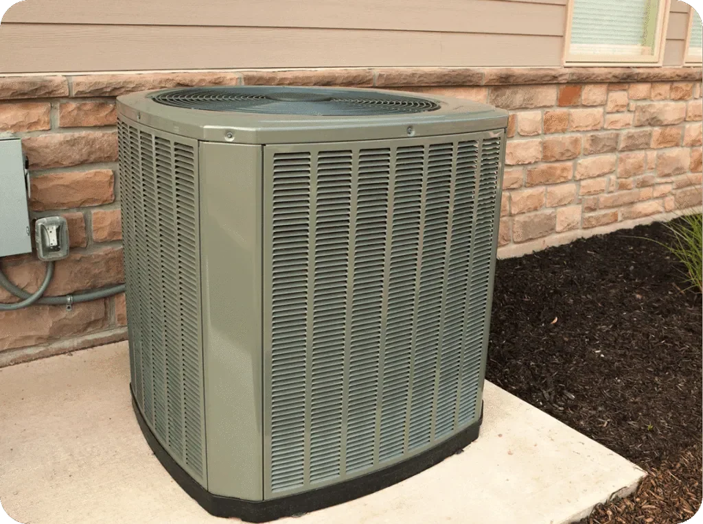 An outdoor air conditioning unit beside a brick house wall and a garden bed with mulch.