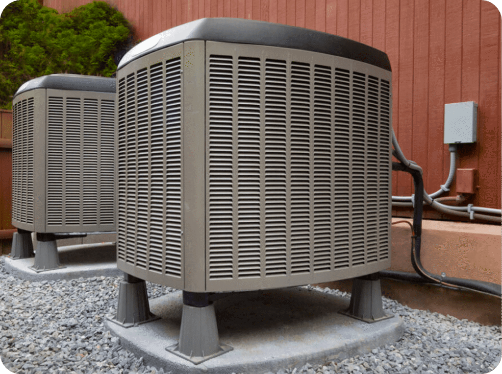 Two outdoor HVAC units on a gravel surface next to a red wooden wall, with electrical wiring and a green bush in the background.