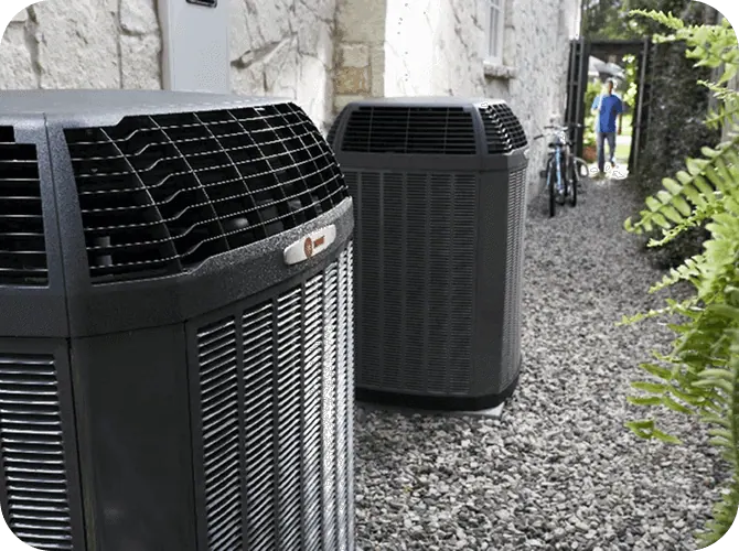 Two large outdoor air conditioning units on a gravel path next to a stone wall, with a person walking a dog in the background.