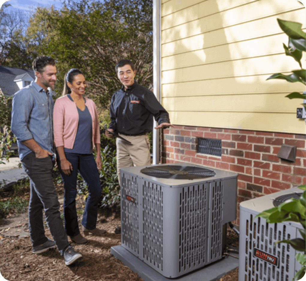A home inspection or consultation taking place outdoors on a sunny day, featuring three adults standing near two HVAC units next to a house with yellow siding and a brick base.