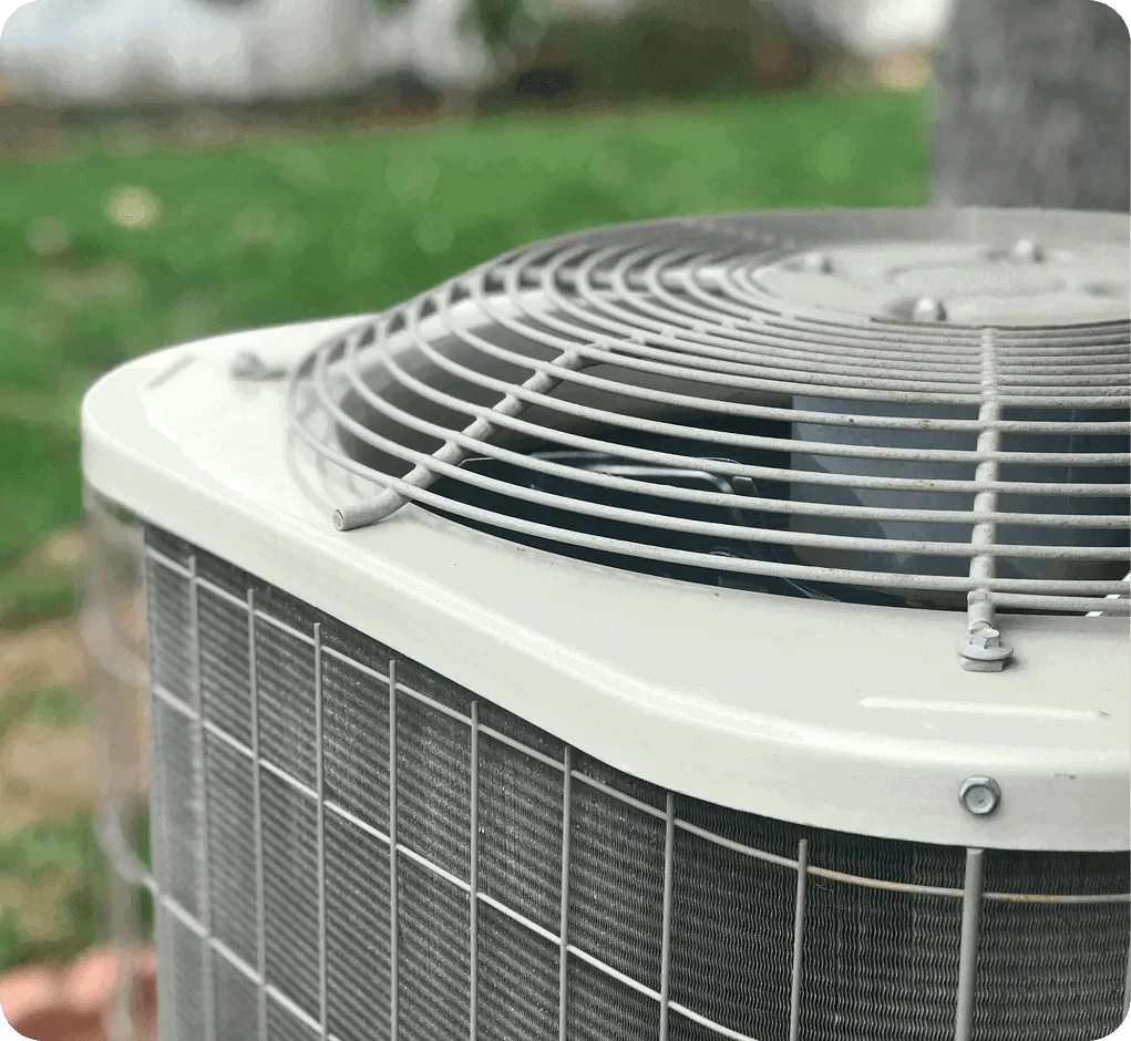 Close-up of an outdoor air conditioning unit with a protective metal grille on top, situated in a backyard with green grass and trees in the background.