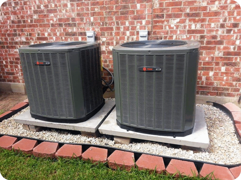 Two air conditioning outdoor units installed outside a brick wall on concrete slabs, surrounded by a border of red bricks and gravel.