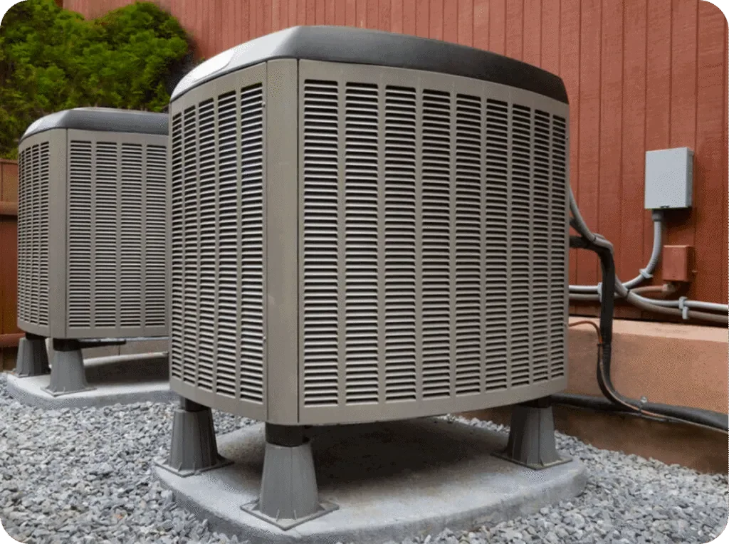 Two outdoor air conditioning units on concrete pads, connected to a building with electrical wires, with a wooden fence and green trees in the background.