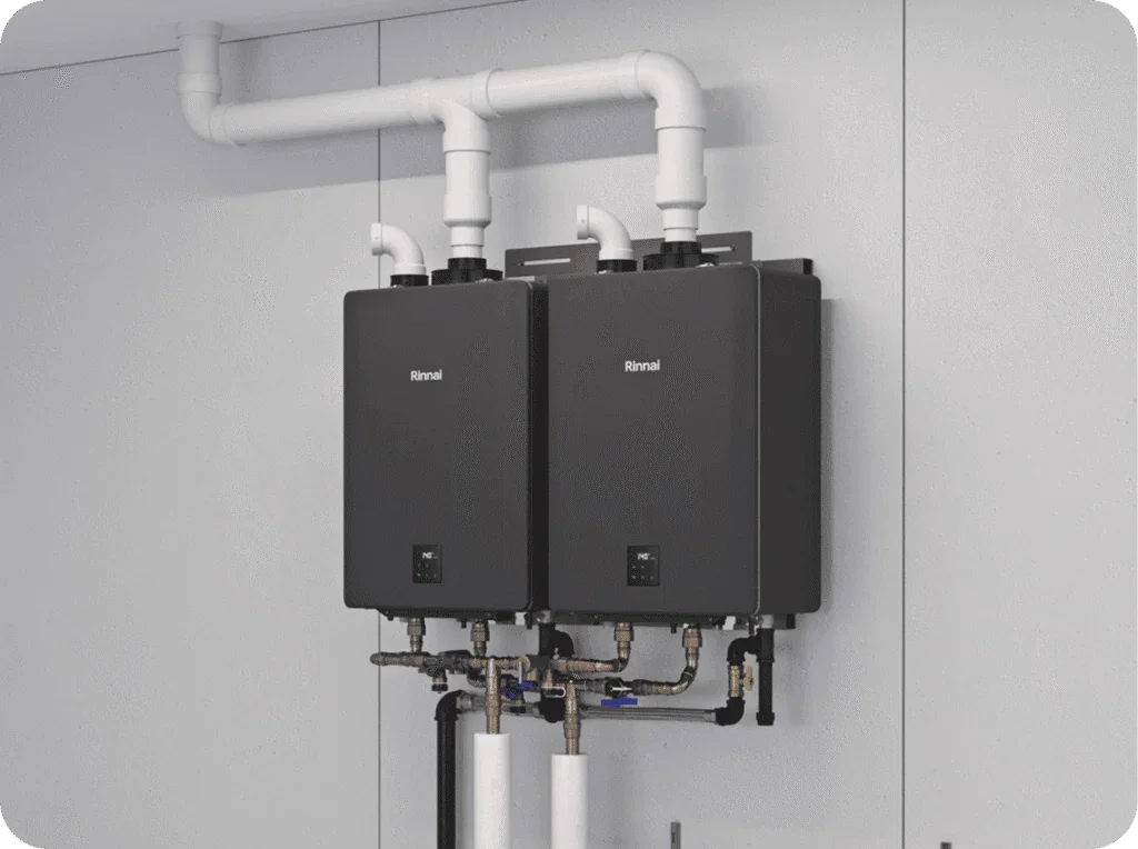 Two black Rinnai tankless water heater units mounted on a gray wall with white PVC pipes connected at the top and various pipes at the bottom.