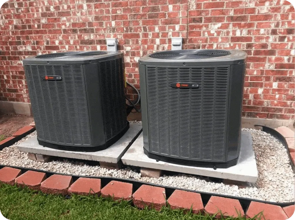 Two HVAC units outside a brick building on concrete slabs with a gravel base, surrounded by red bricks and green grass.