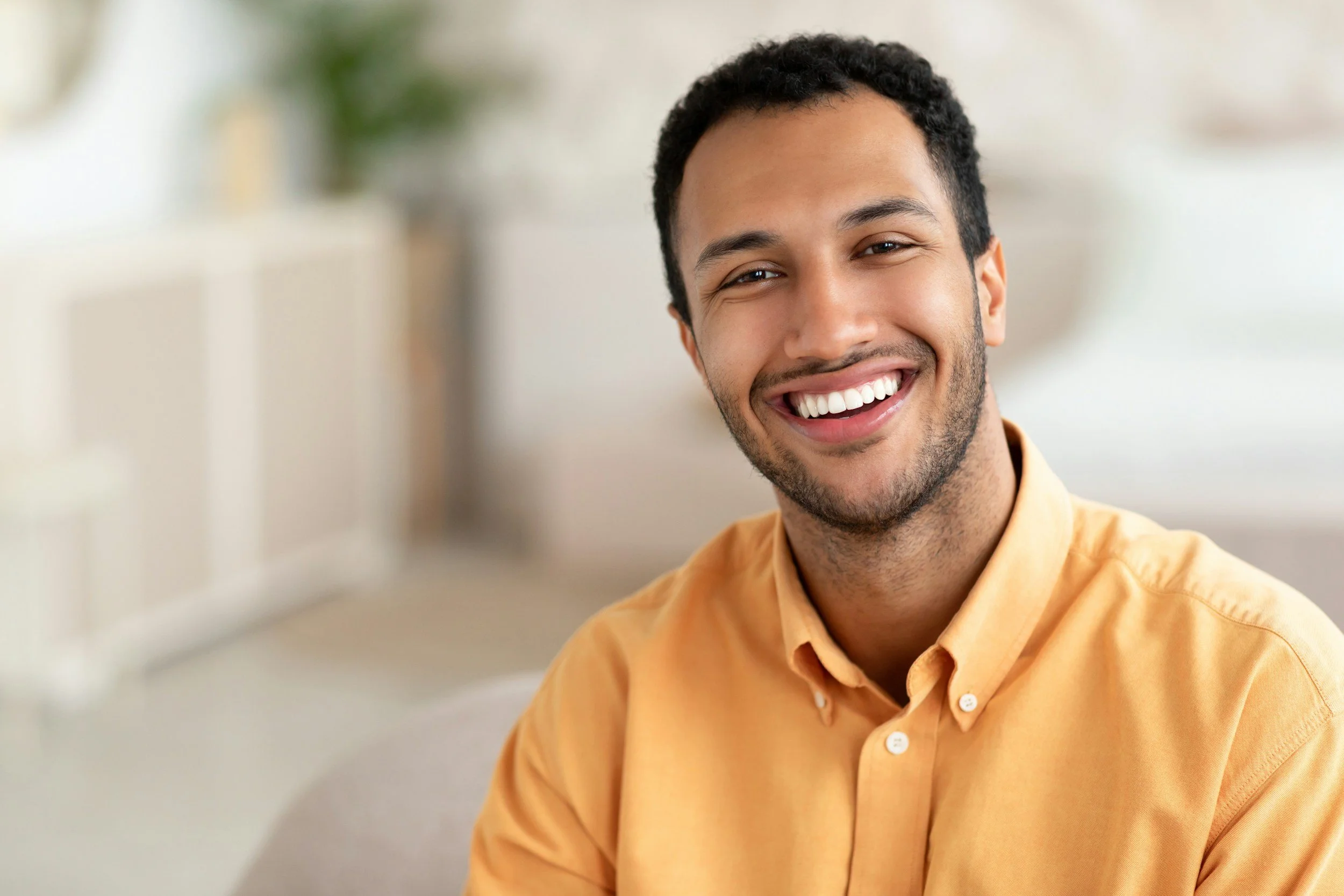 A smiling man with dark curly hair, facial hair, wearing a light orange button-up shirt, indoor background.