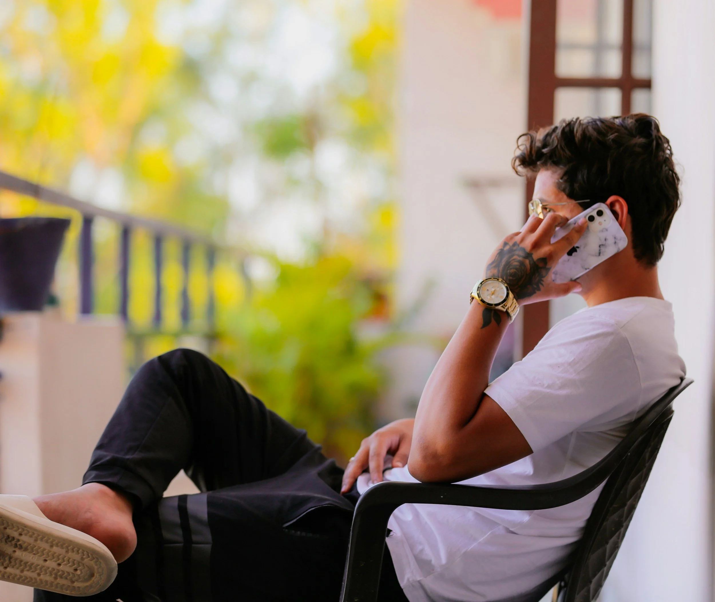 Young man with tattoos, glasses, and a gold wristwatch talking on a cell phone while sitting in a chair, with another person whose legs are visible in the foreground.