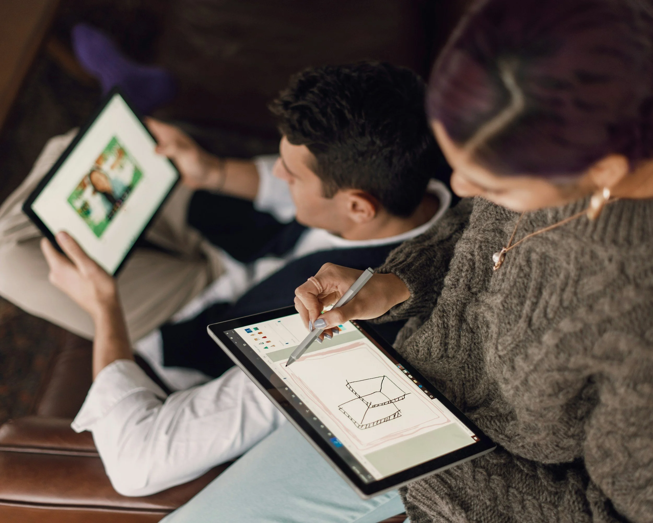 Two individuals sitting side by side; one person is drawing a 3D shape on a tablet with a stylus, while the other person appears to be observing or taking notes. The scene suggests a collaborative or educational setting focused on design or technical drawing.