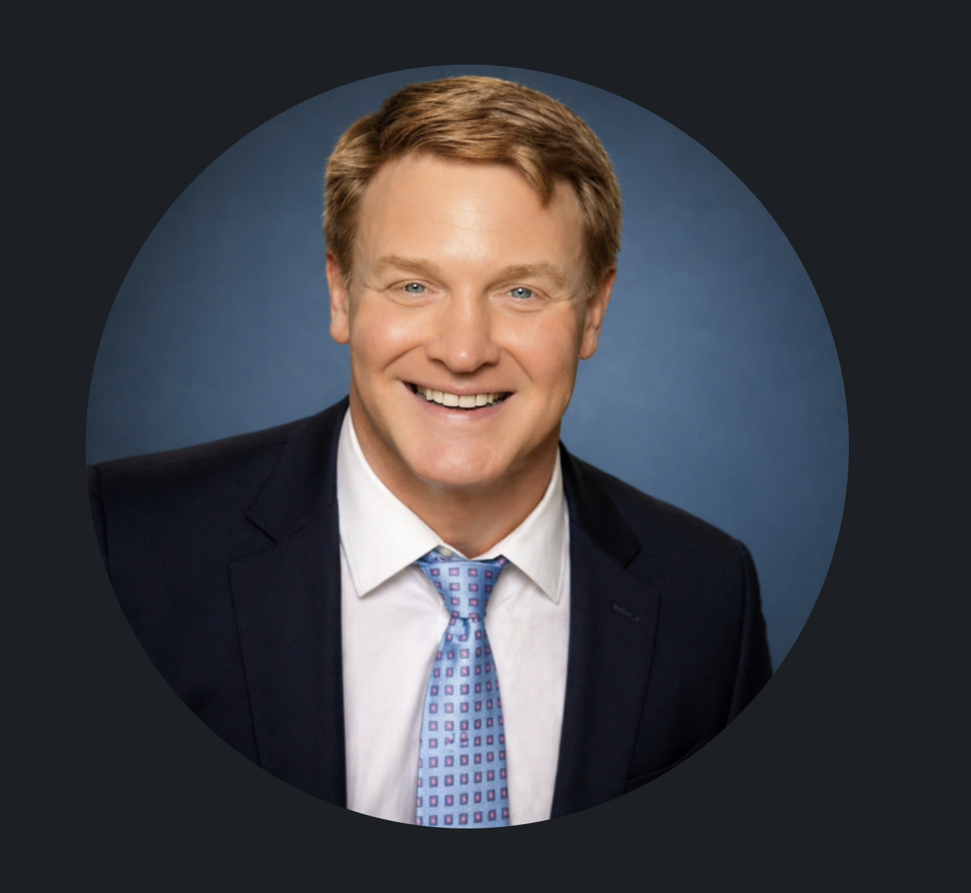 Headshot of a man with short red hair smiling, wearing a dark suit, white shirt, and a blue patterned tie, against a blue background.