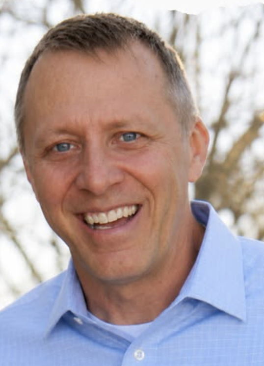 Close-up of a smiling man with short brown hair and blue eyes, outdoors with blurred trees in the background, wearing a light blue collared shirt.