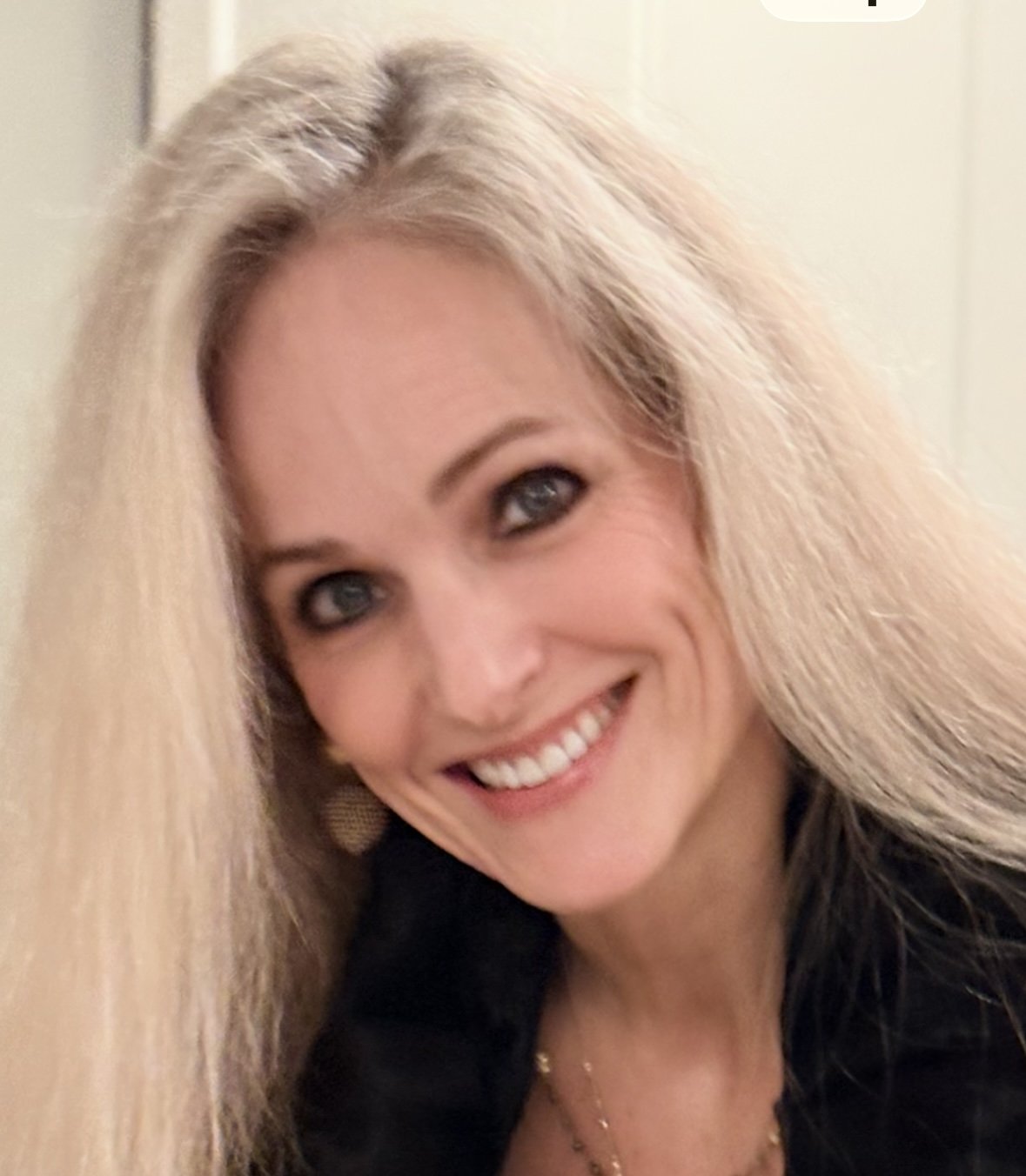 A smiling woman with long blonde hair, blue eyes, wearing earrings and a black top, posing indoors.