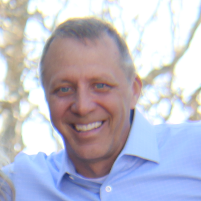 A smiling man wearing a light blue dress shirt outdoors with trees in the background.