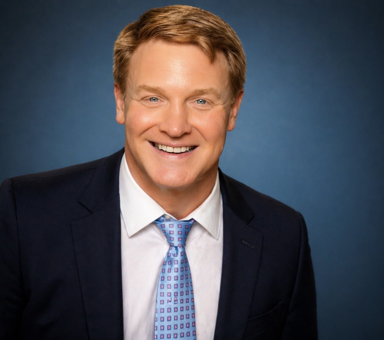 Headshot of a smiling man in a dark suit, white shirt, and patterned tie against a blue background.