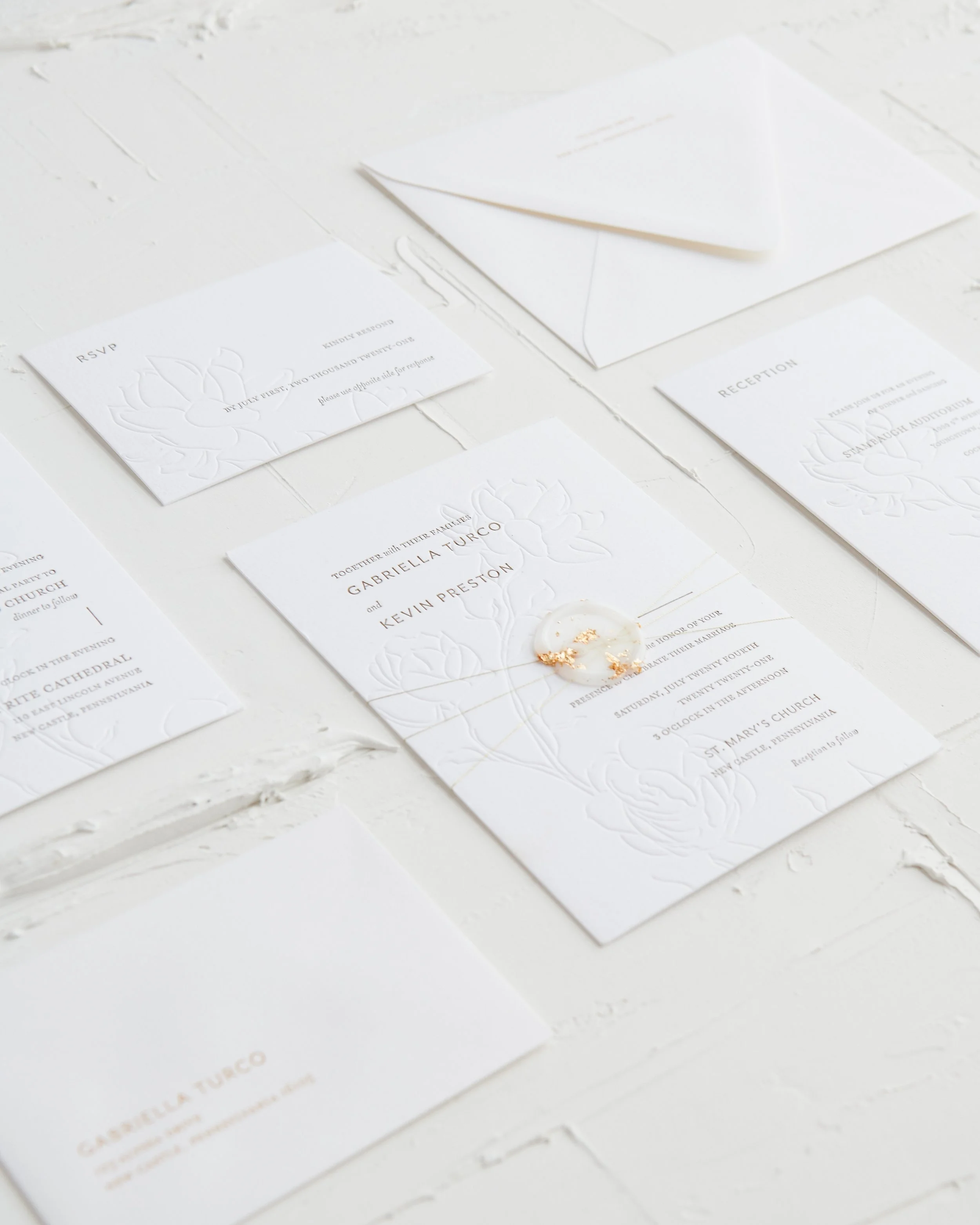 Wedding invitation suite including RSVP card, reception card, and menu card, all with embossed floral design and gold accents on white paper, laid out on a white textured surface.