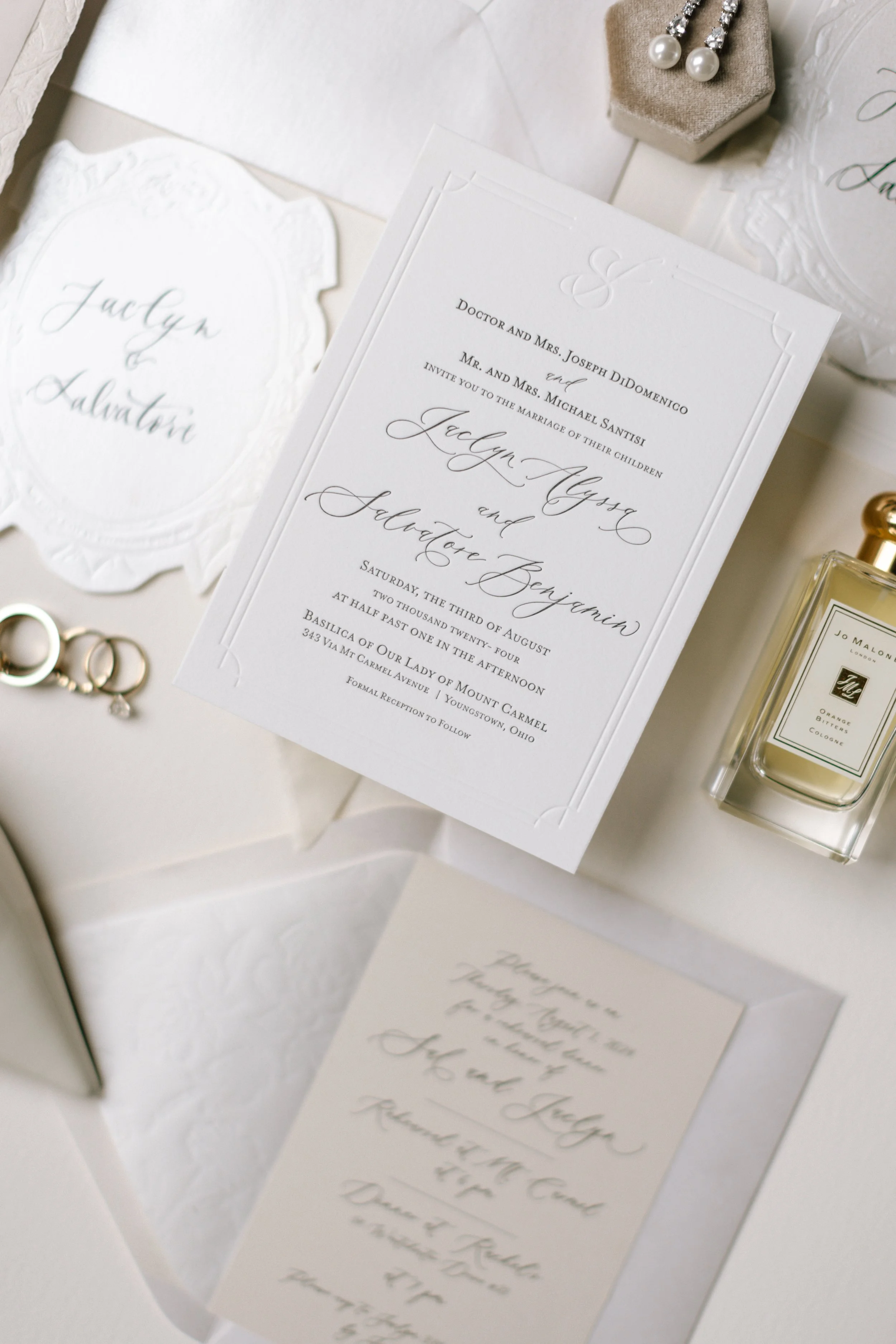 Wedding invitation with elegant calligraphy, pearl earrings on a beige jewelry stand, and a bottle of perfume on a white surface.