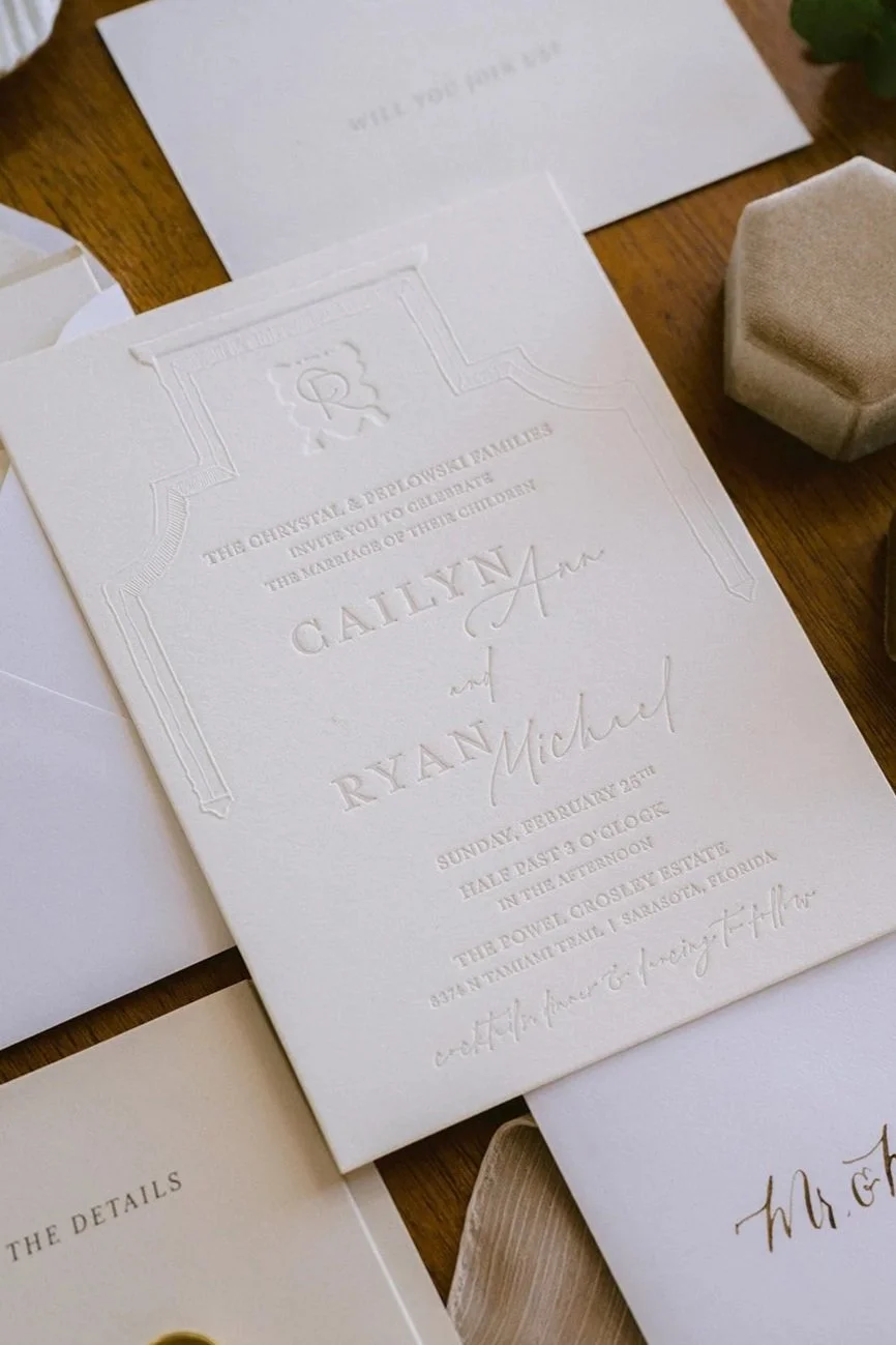 Elegant wedding invitation on white textured card stock with embossed details, announcing the marriage of Cailyn and Ryan Michael, with wedding details in small print. Surrounded by other wedding stationery and a small beige box on a wooden surface.